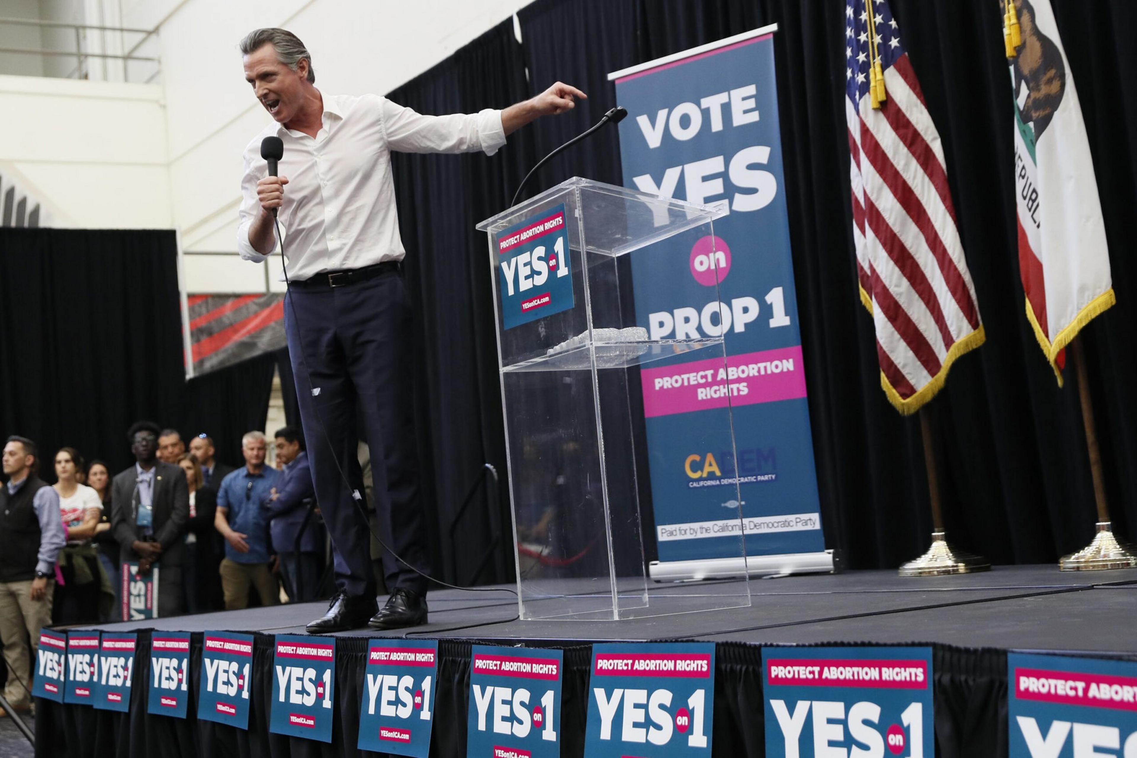A man in a white shirt and dark pants speaks passionately into a microphone on stage with banners reading "YES on PROP 1" supporting abortion rights.