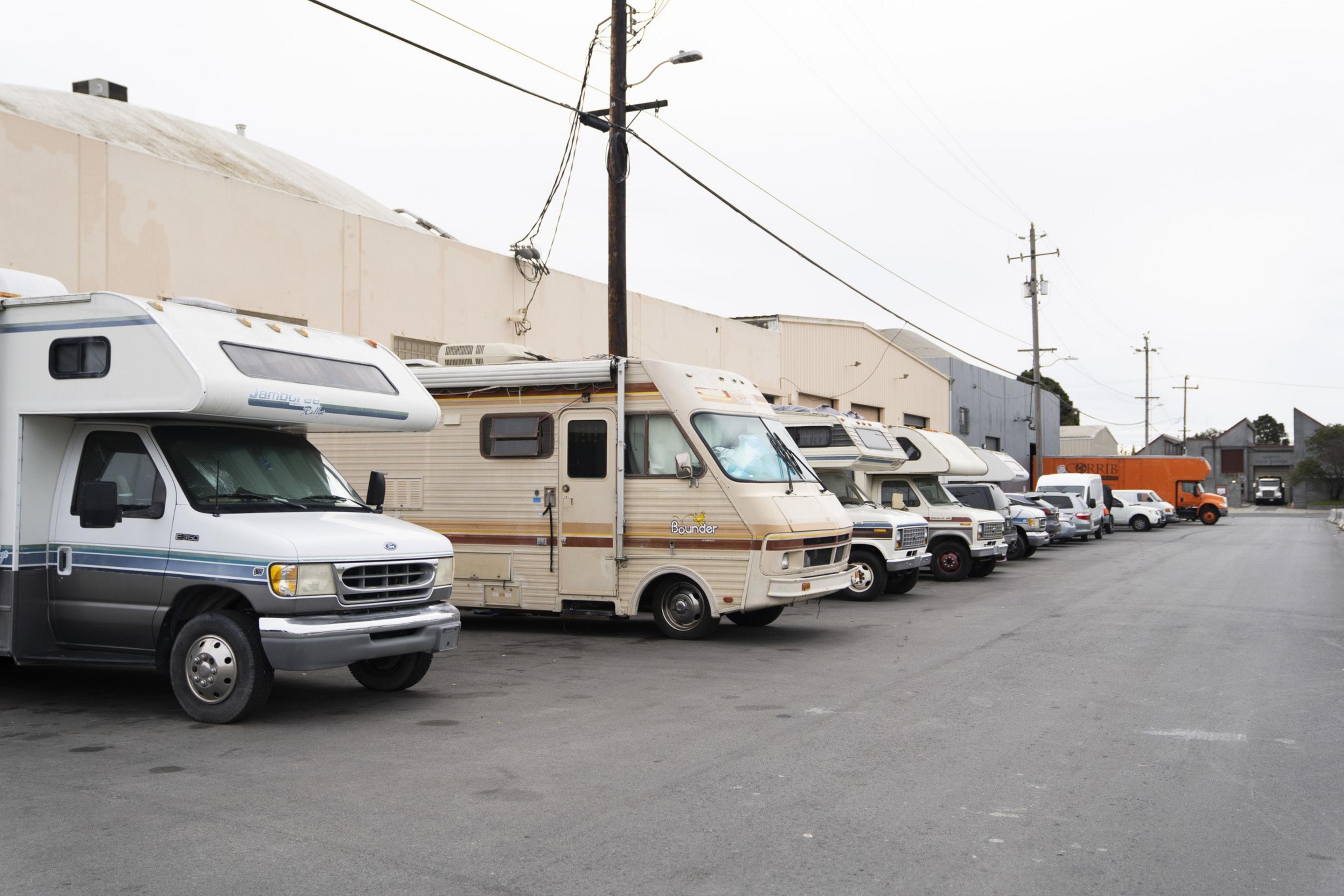 Several RVs are parked along a quiet urban street beside industrial buildings, with power lines overhead and a few cars nearby.