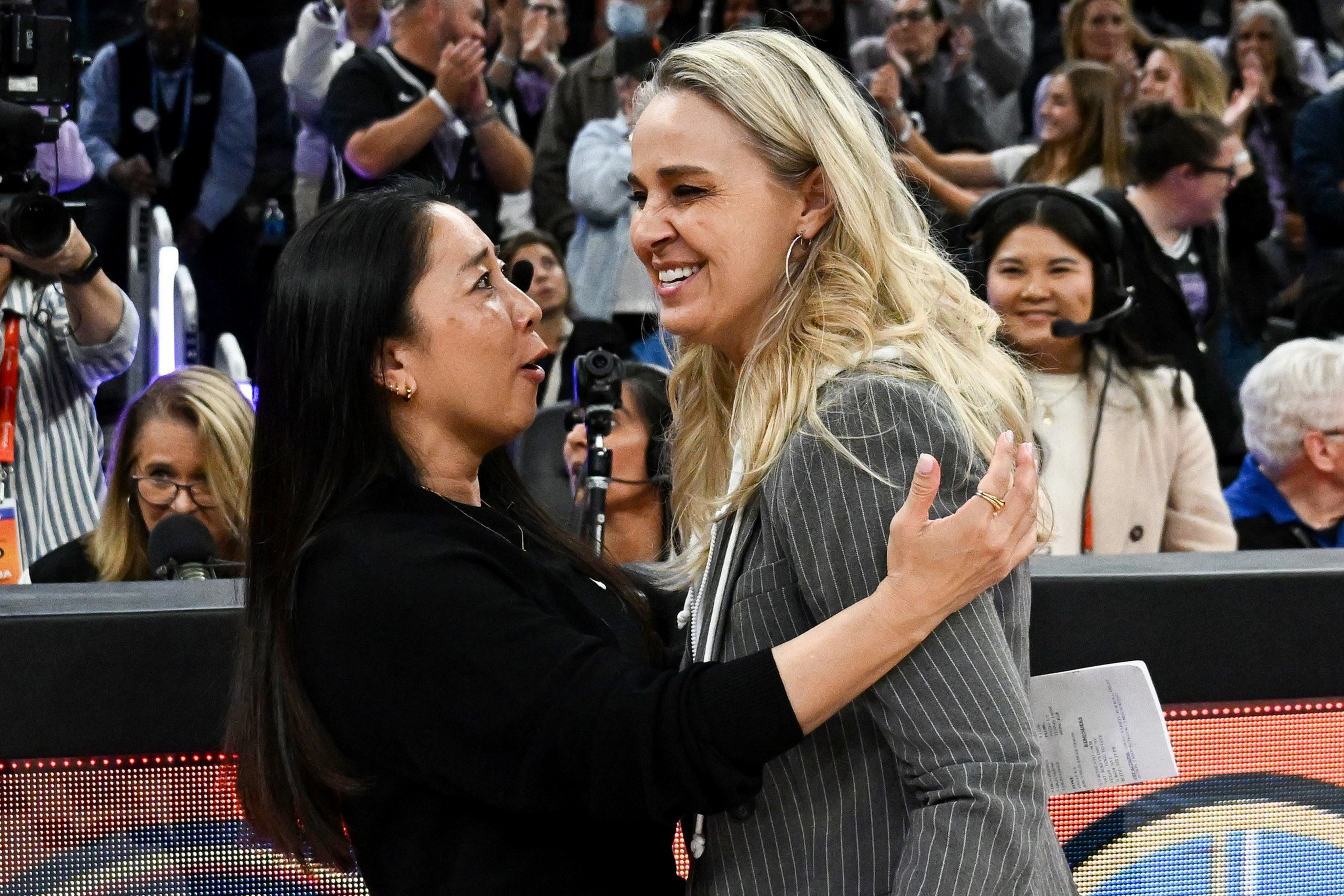Two women warmly embrace and smile, surrounded by a crowd of onlookers and media personnel in a lively indoor setting.