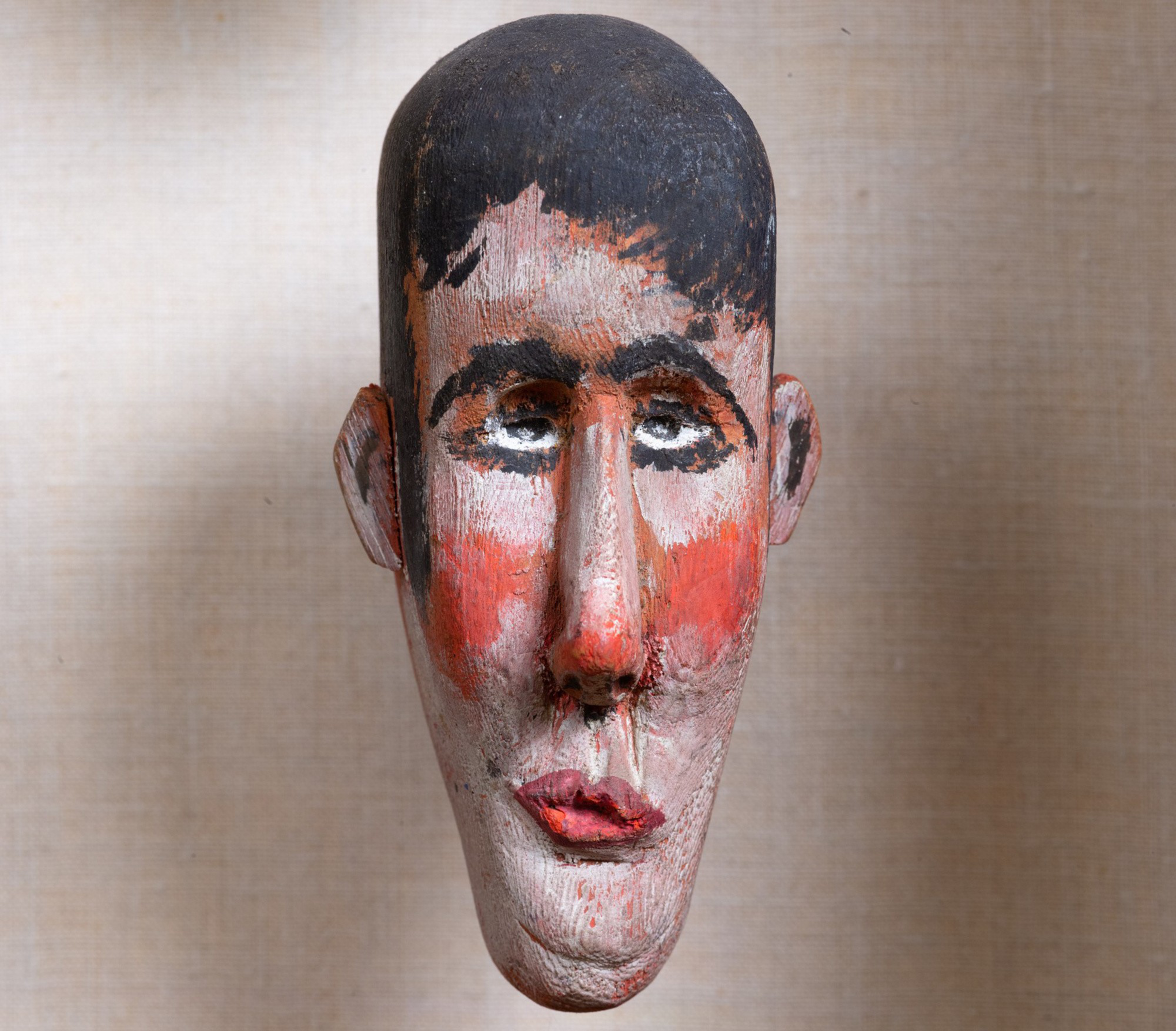 A wooden carved face painted with dark hair, red cheeks, pronounced nose, lips, and black eyebrows against a neutral fabric background.