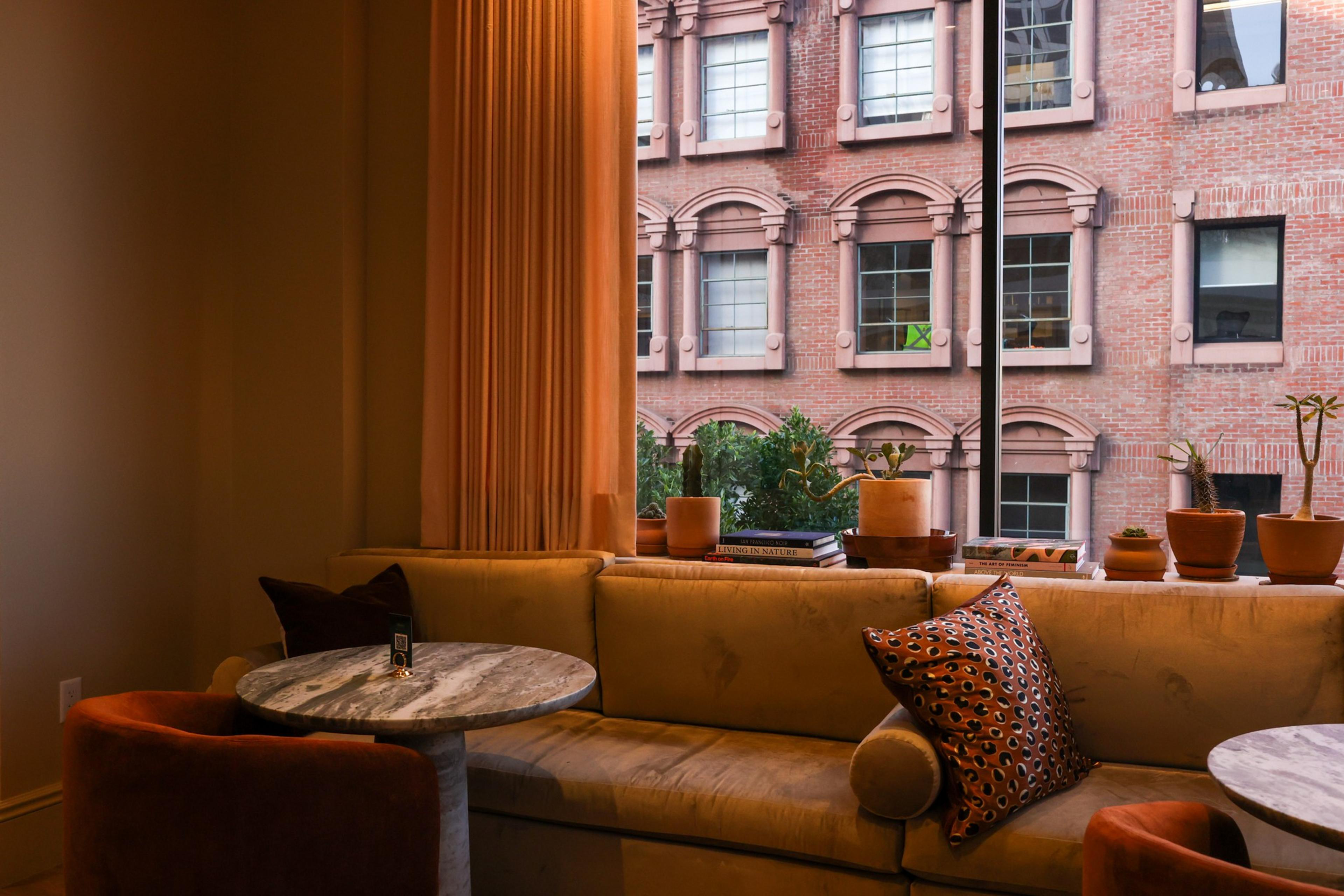 A cozy seating area with a beige couch, a patterned pillow, round marble tables, potted plants on the windowsill, and a brick building visible outside.