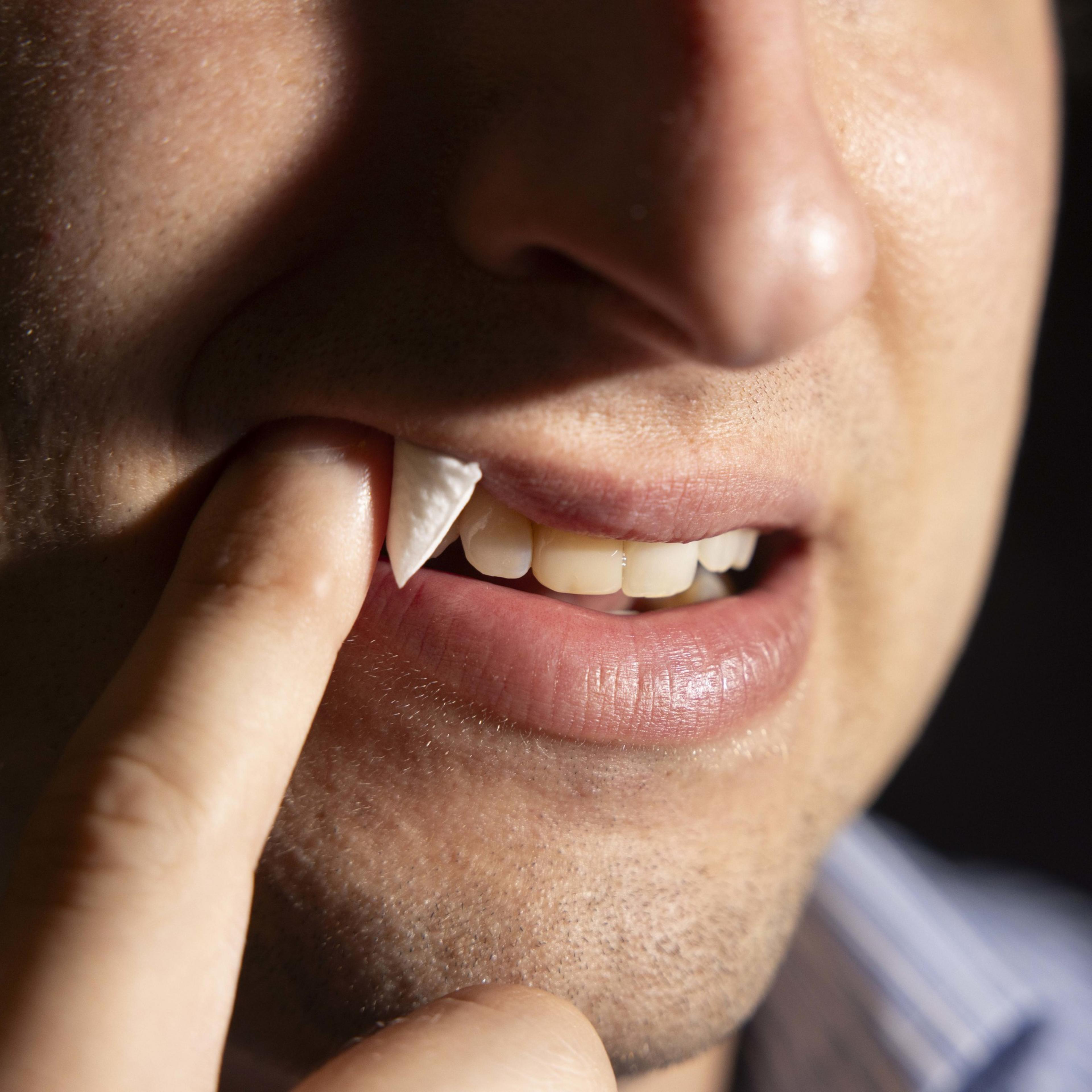 A close-up shows a person pushing a piece of white paper between their teeth and lip with their finger, focusing on their half-smiling mouth and clean teeth.