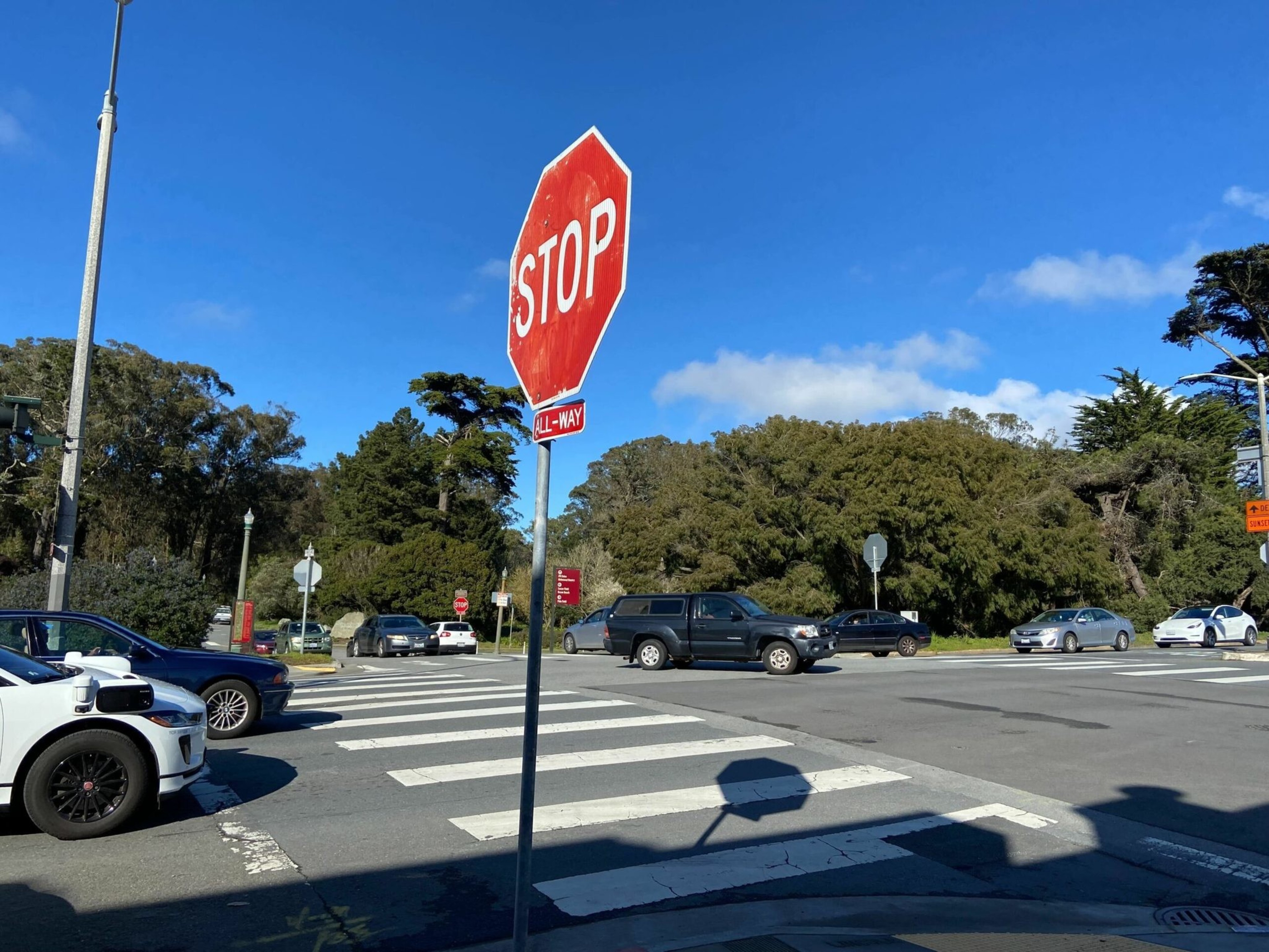 A busy intersection with multiple cars, a crosswalk, a red octagonal stop sign, and trees under a clear blue sky.