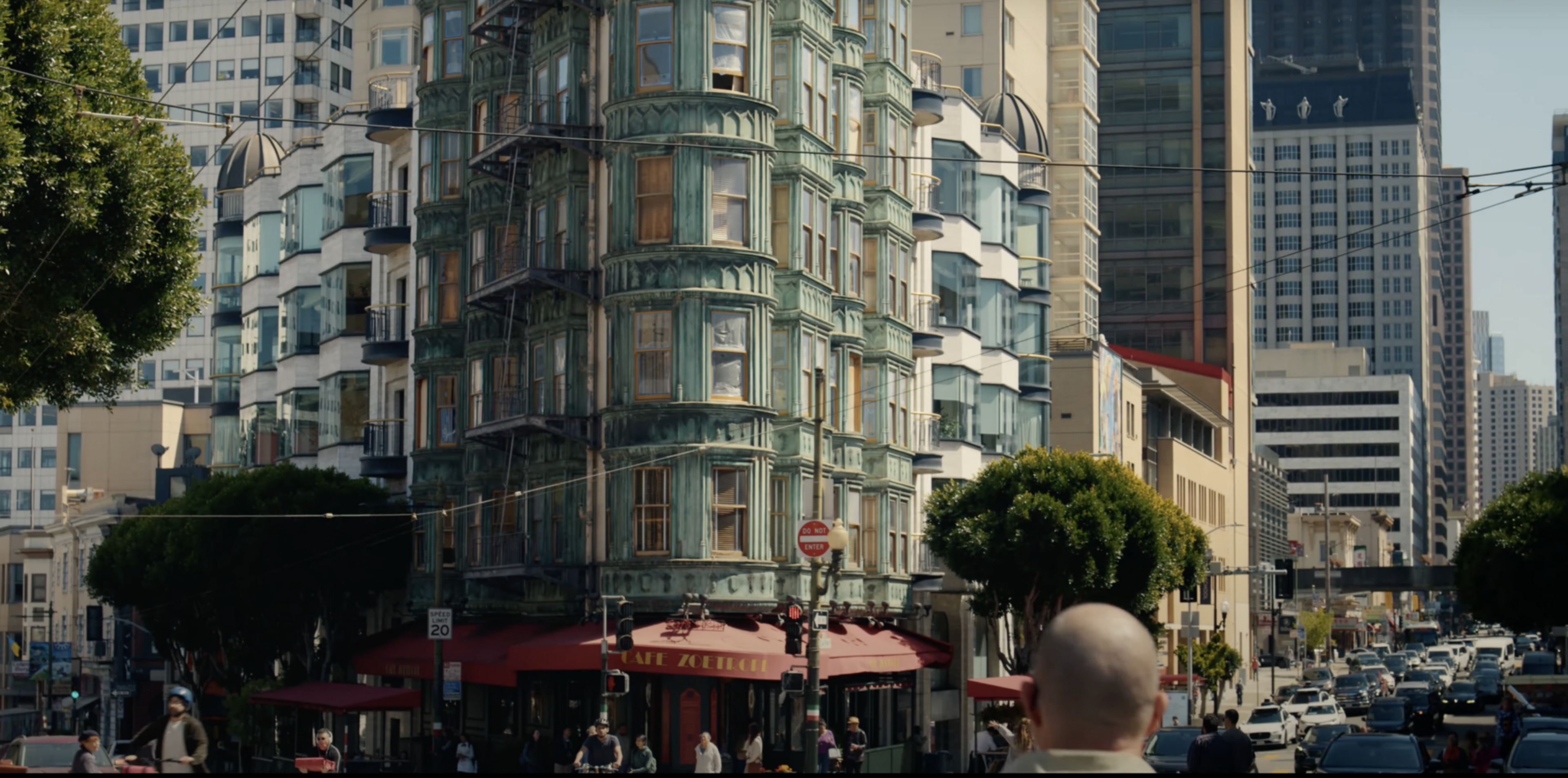 A bustling city street with a green ornate building at an intersection. People walk near a cafe with a red awning, surrounded by taller modern buildings.