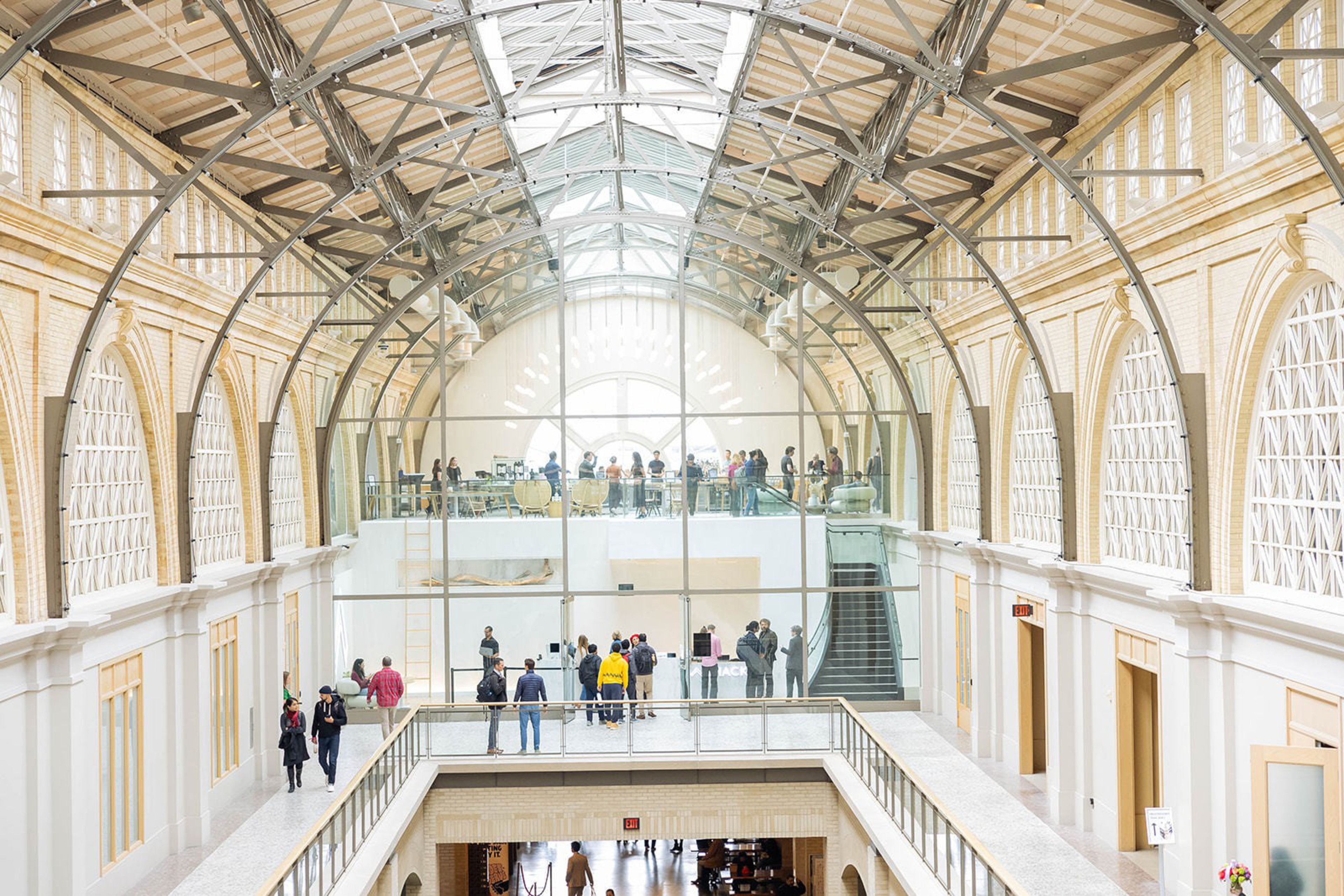 The image shows a spacious, bright indoor area with a high, arched glass and steel ceiling. People are gathered on a mezzanine level, and more are seen below.