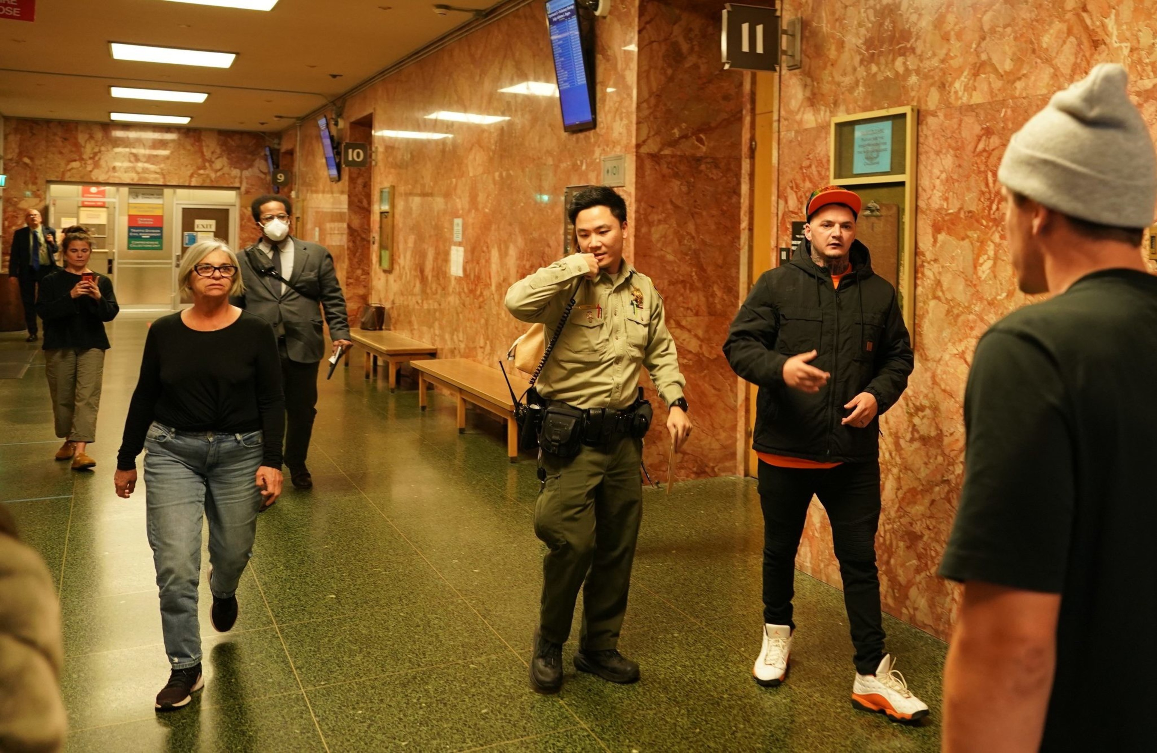 People walk through a hallway with marble walls and a green floor. One person is a uniformed officer, and others appear to be casually dressed. An exit sign is visible.