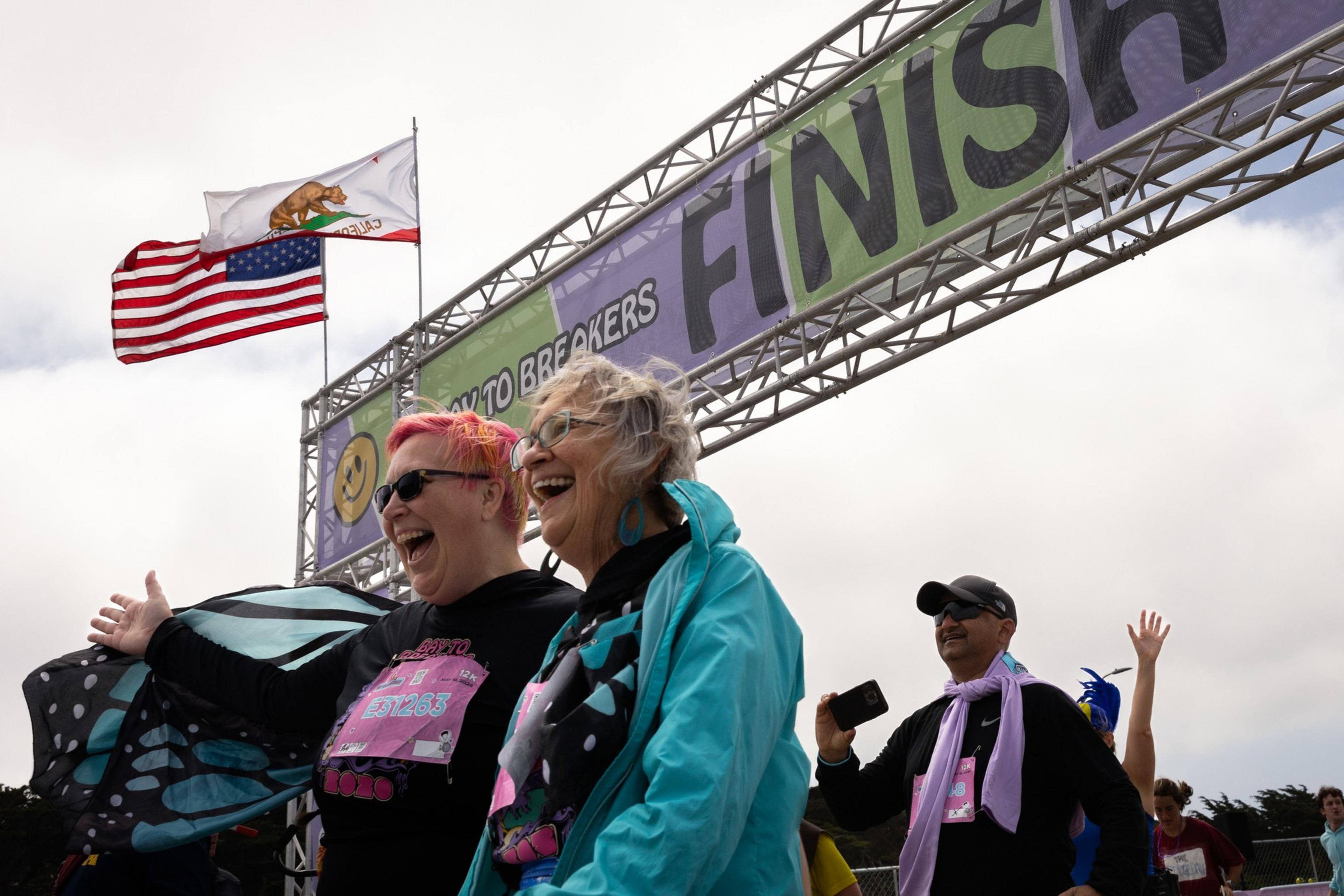 Three joyful people are at a race finish line under a banner that reads &quot;FINISH&quot;; one waves a butterfly-shaped kite, another laughs beside her, and a man records the moment. Flags flutter above.