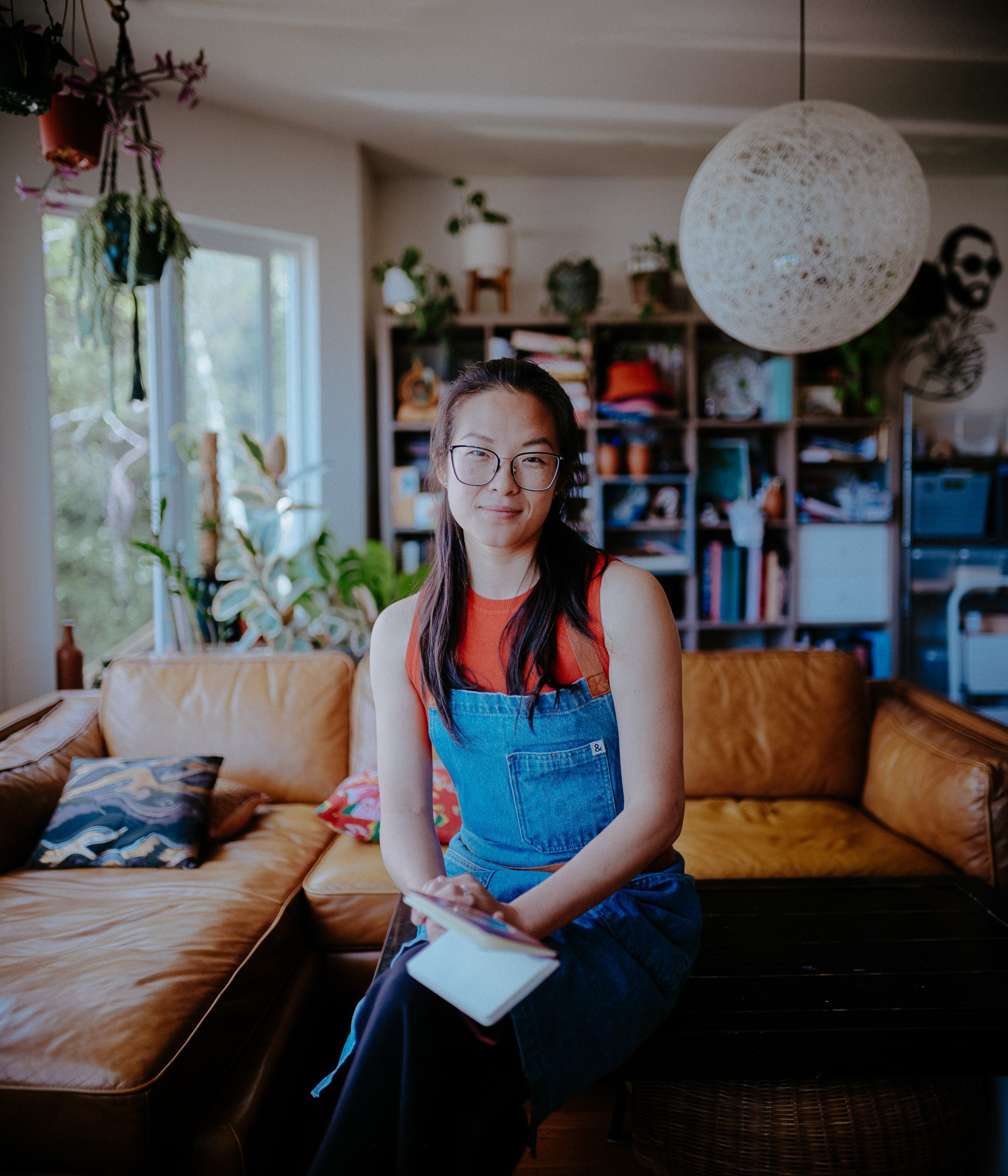 A woman with glasses and long hair sits on a brown leather sofa, holding a notebook in a cozy room filled with plants and shelves.