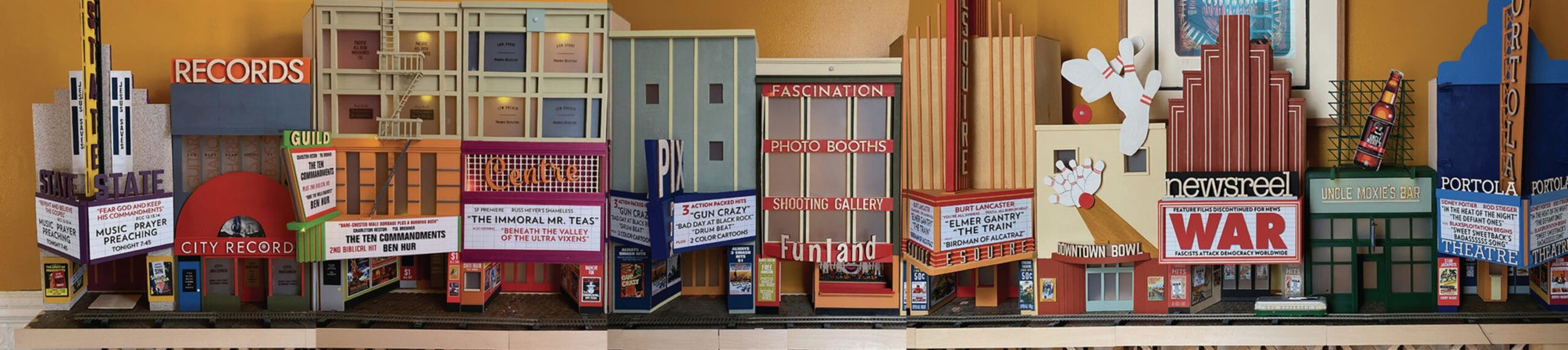 A detailed miniature cityscape features theater marquees, record signs, and colorful billboards with names like "CITY RECORD," "WAR," and "Funland."