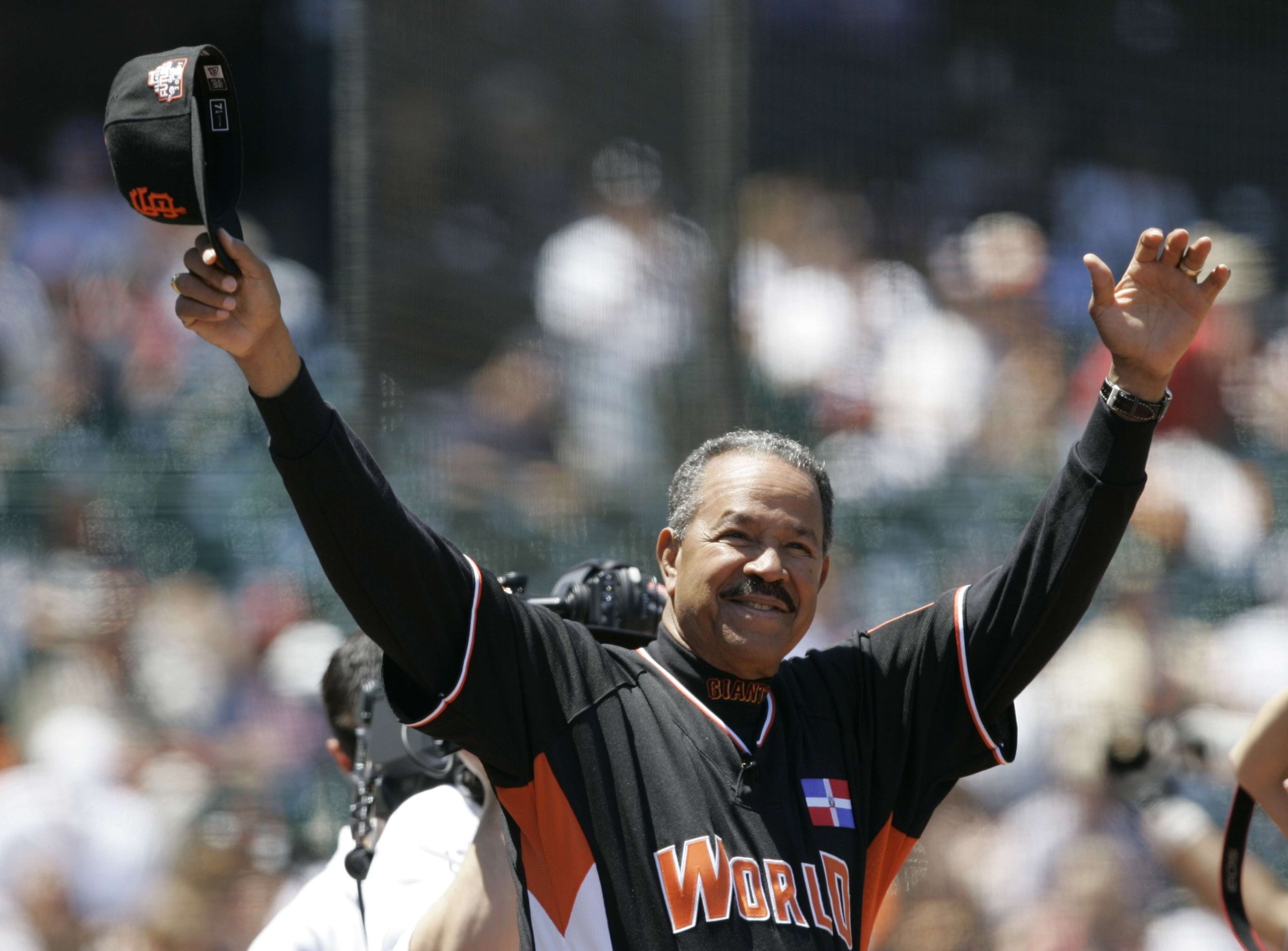 A smiling man in a &quot;World&quot; baseball jersey raises his arms, holding a cap in one hand, with a camera around his neck. The background shows a blurry crowd.