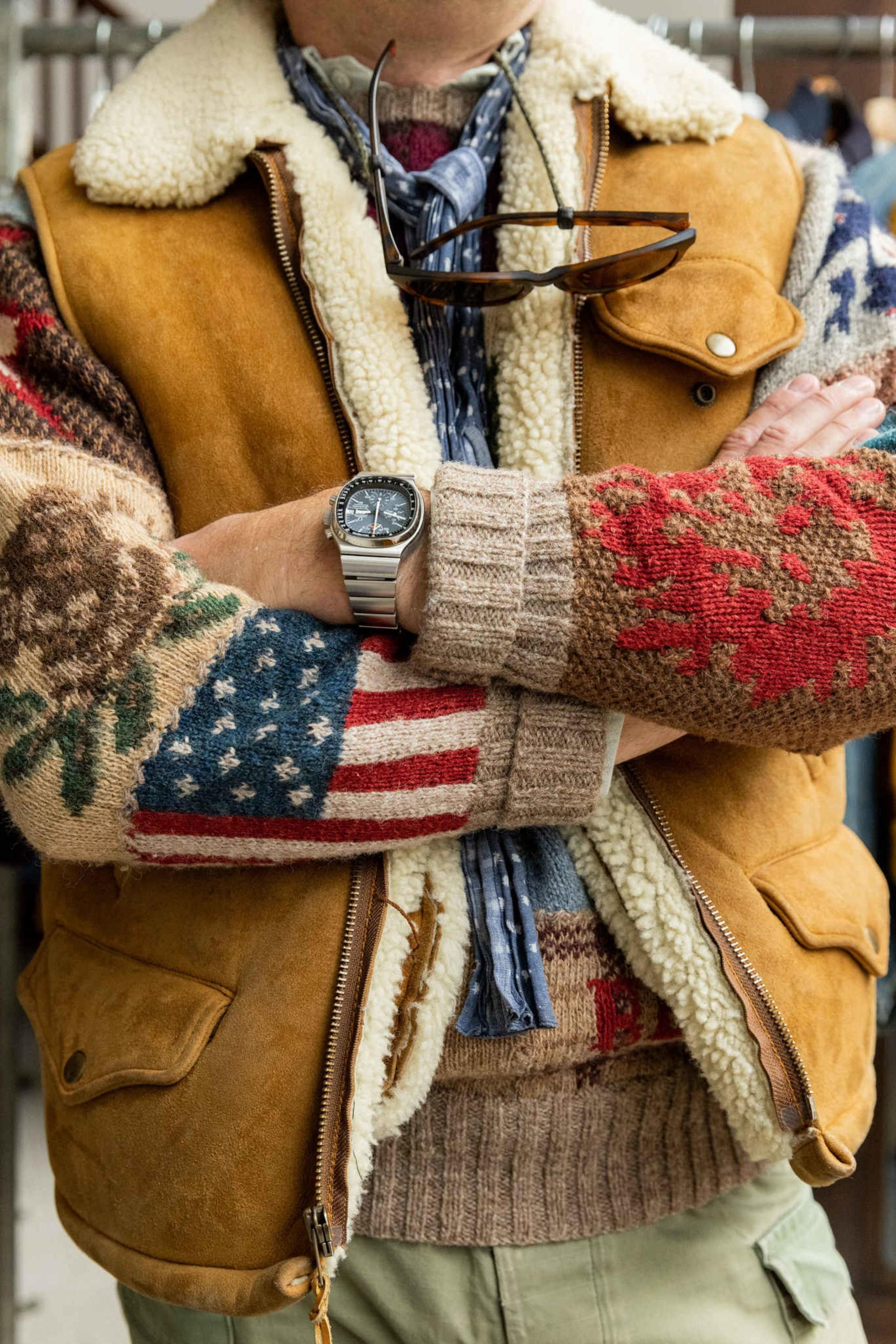 A person wears a colorful patterned knit sweater with an American flag sleeve, a brown shearling vest, sunglasses hanging from a scarf, and a silver watch.