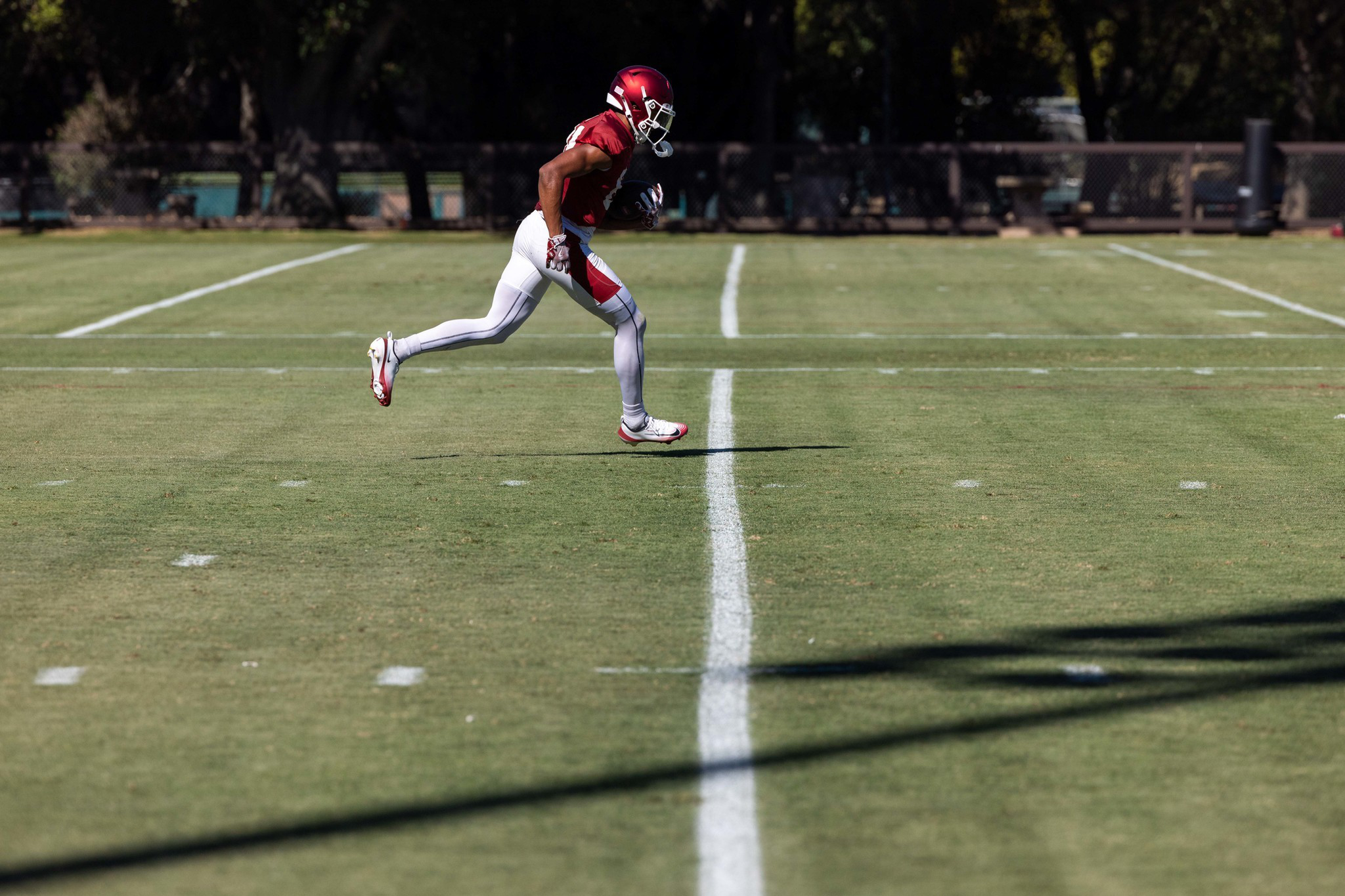 A football player in red and white uniform runs across a grassy field while holding a football tightly under one arm.