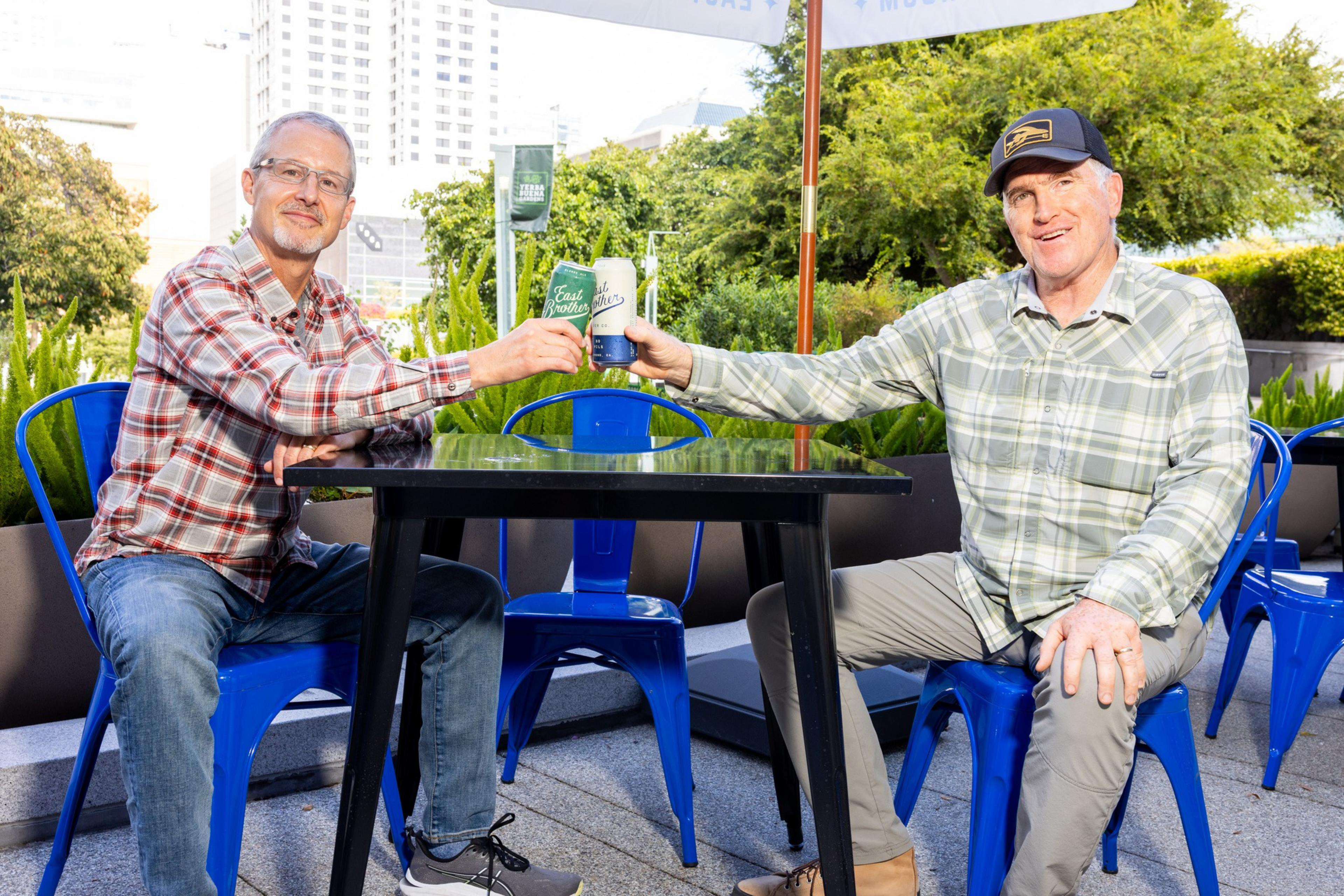 Two men in plaid shirts sit on blue chairs at a black table outdoors, clinking beer cans and smiling with greenery and buildings in the background.