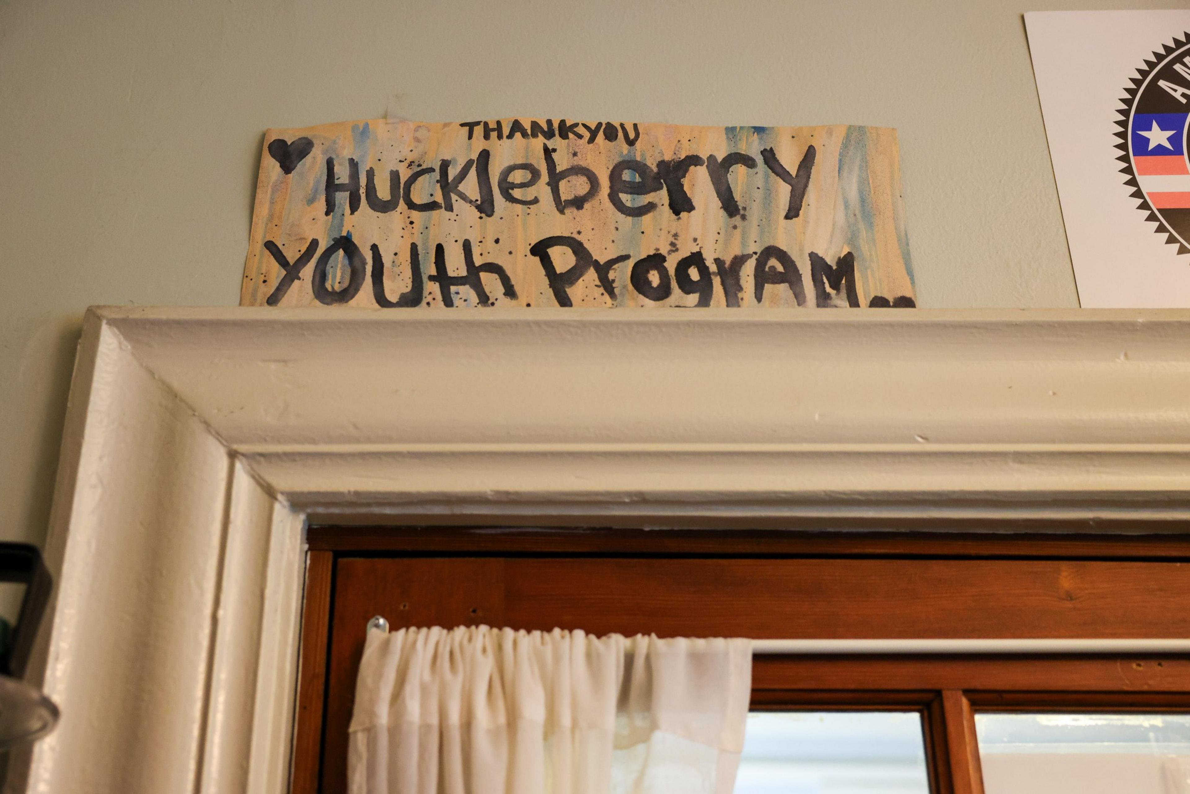 A hand-pained butter yellow and light blue sign reading &quot;Thank You Huckleberry Youth Program&quot; sits on the cream molding above a brown wood French door.