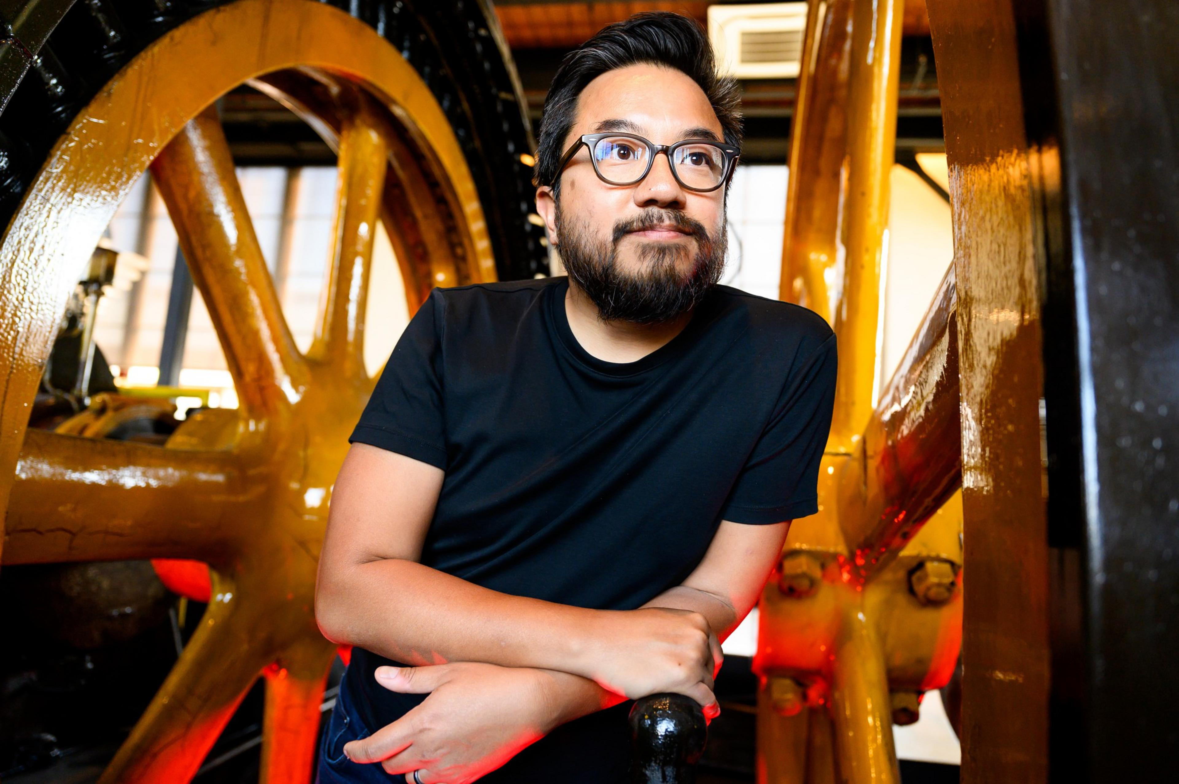 A man with glasses leaning on a large, industrial wheel with a thoughtful expression.