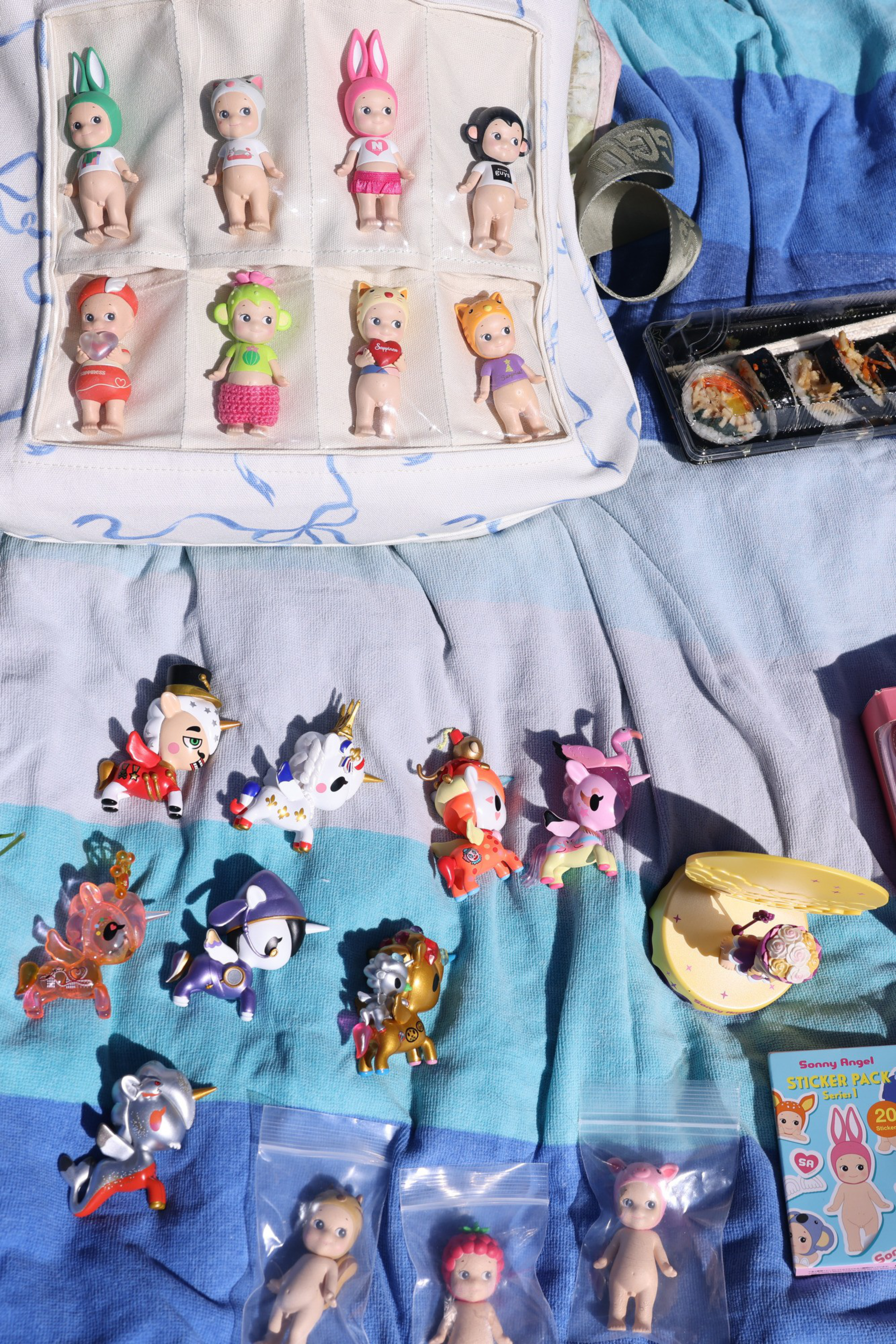 The image shows various small figurines, including ones with animal hats, and colorful unicorns on a striped cloth. There's sushi and a sticker pack nearby.
