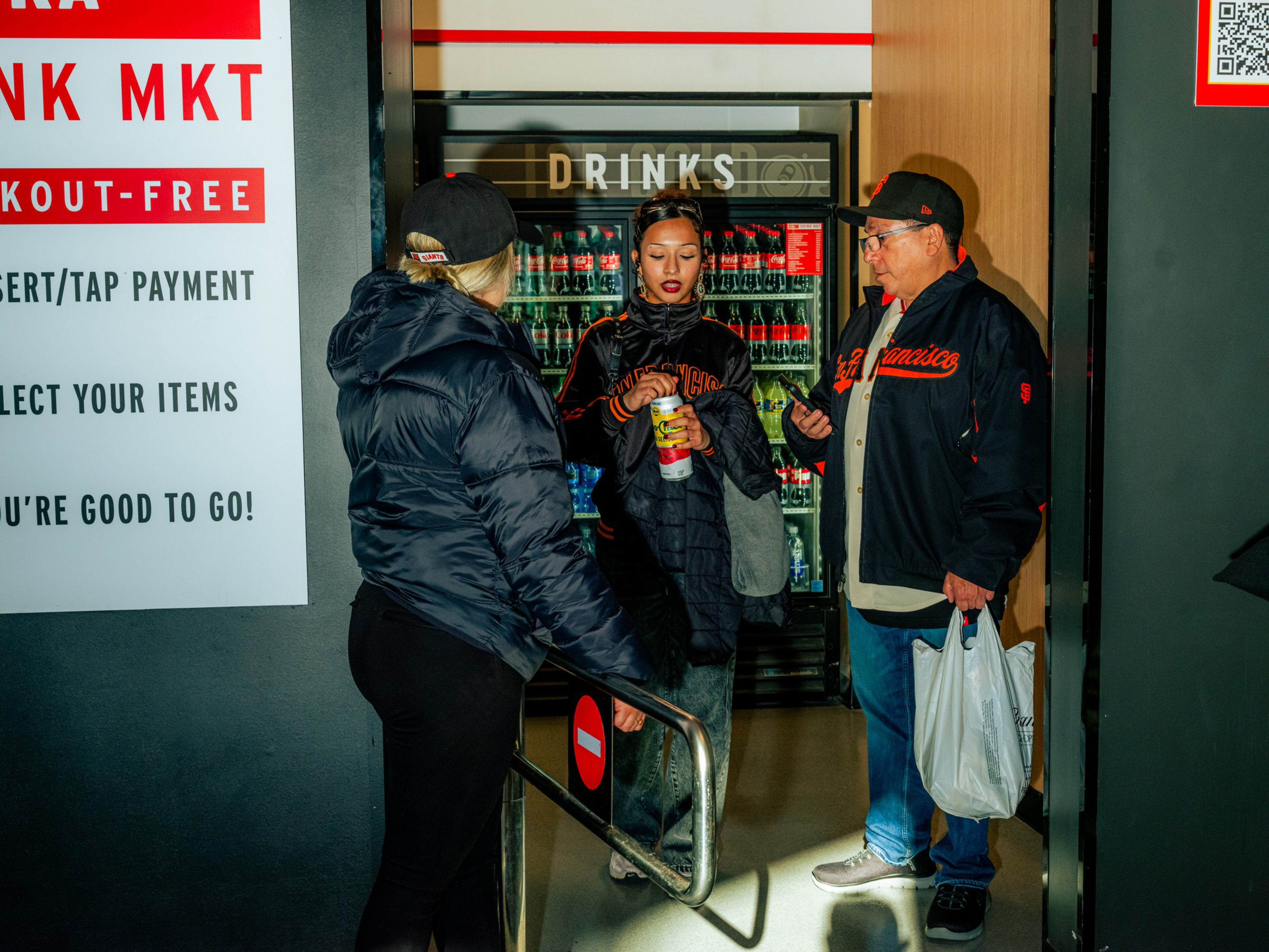 Three people stand near a drink cooler; one holds a can, another carries a shopping bag, and a woman in a black jacket opens a gate marked with a no-entry sign.