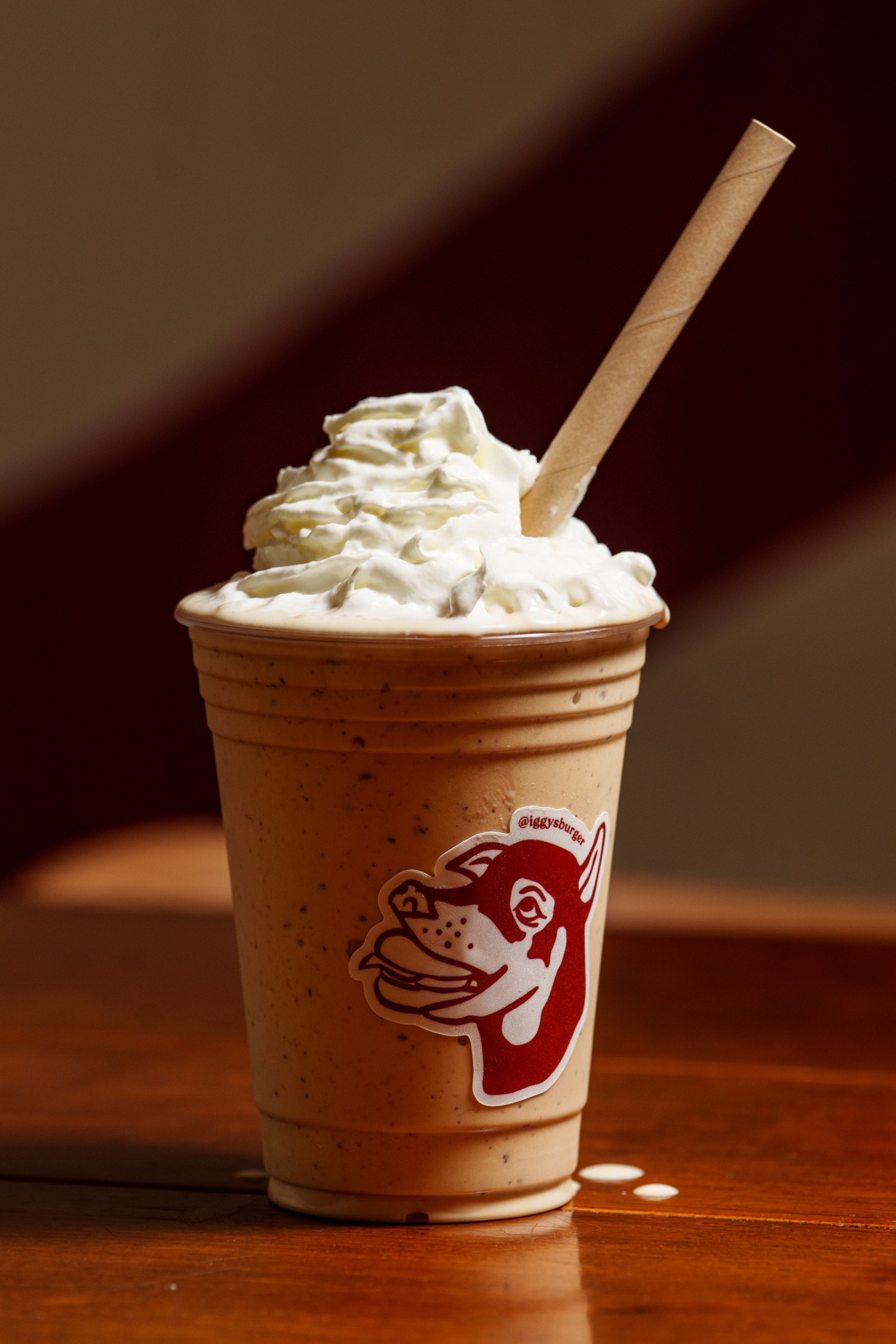 A creamy milkshake with whipped cream on top is in a plastic cup featuring a stylized dog face logo, with a paper straw inserted.