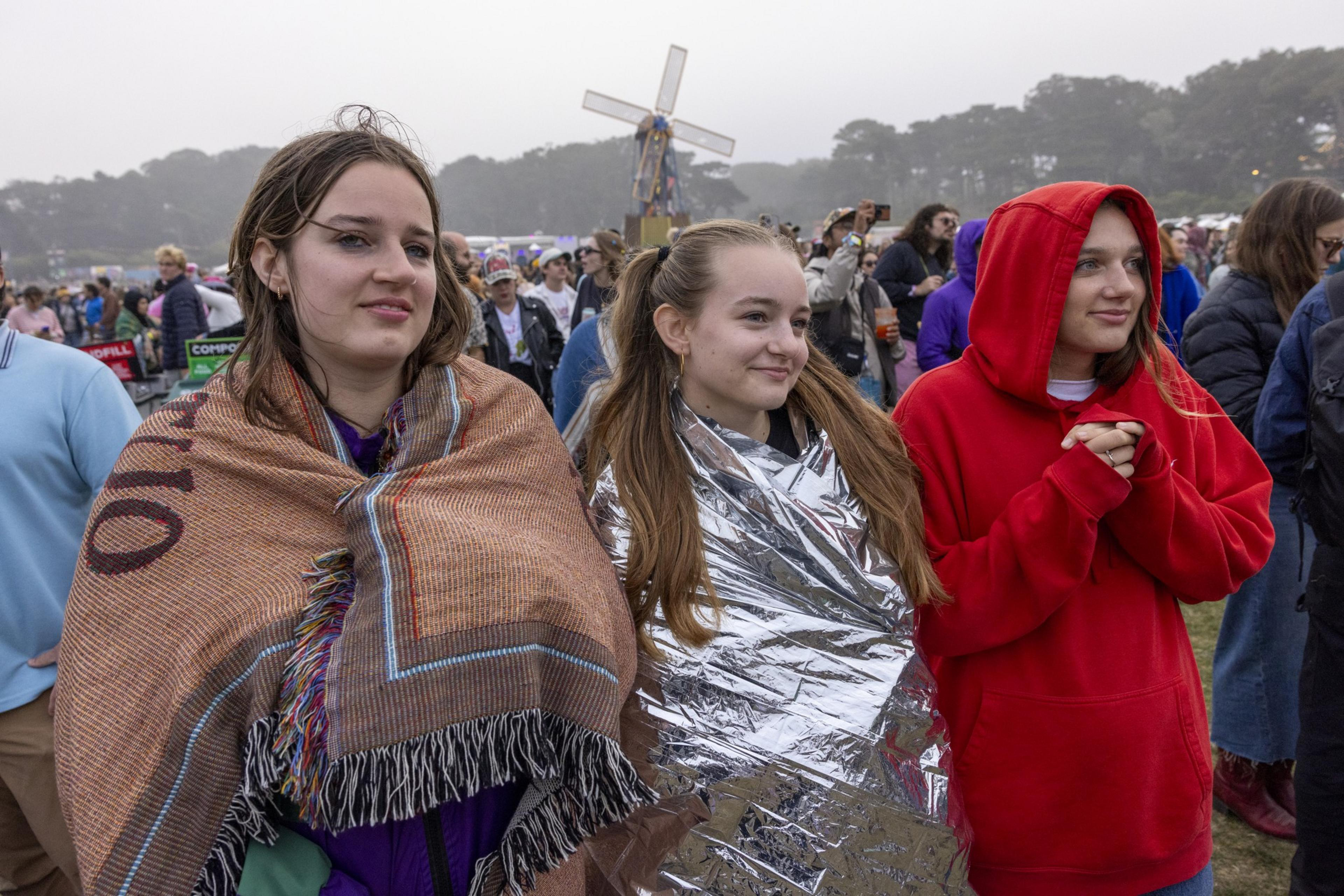 Three young women are wrapped in blankets and a jacket, standing in a crowd outdoors. One wears a brown blanket, another a foil one, and the third a red hoodie.