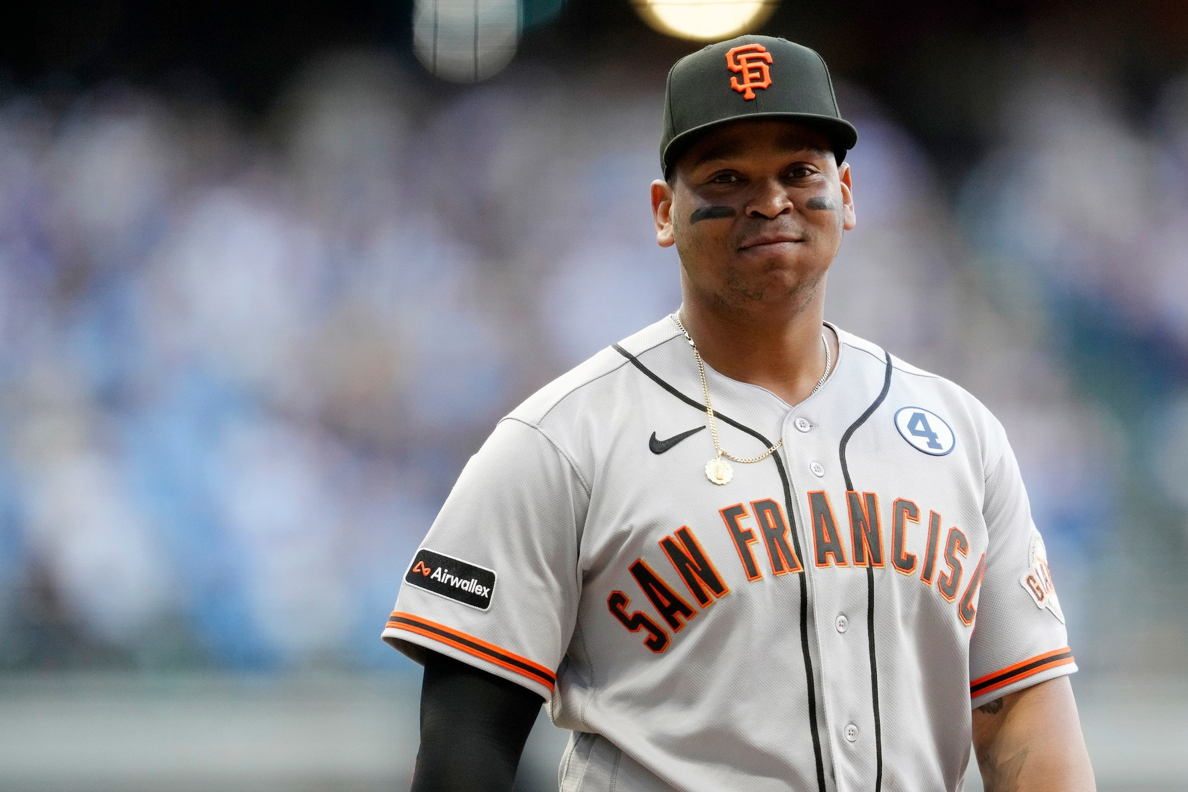 A baseball player in a San Francisco Giants gray jersey and black cap stands with a slight smile, wearing eye black and a gold necklace.