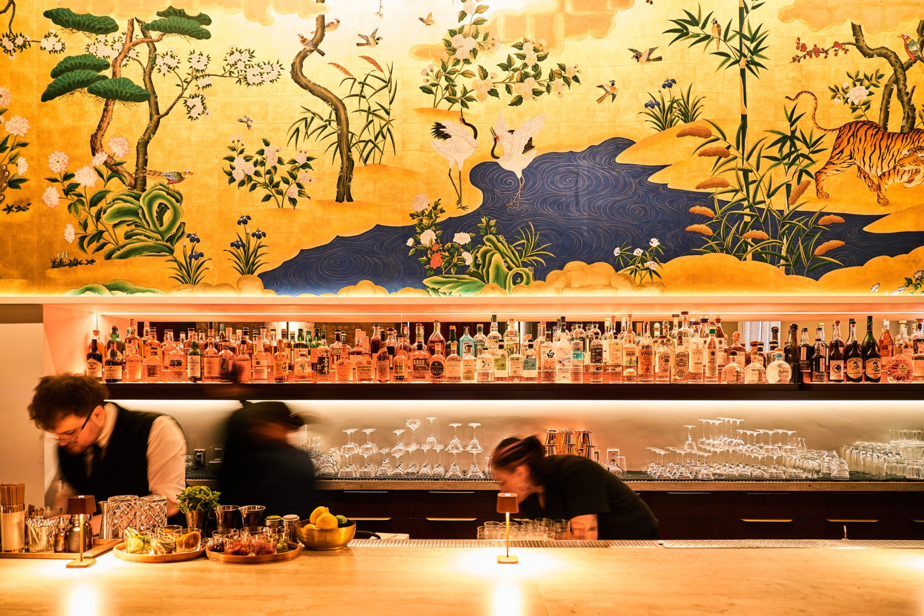 A vibrant bar scene features three bartenders preparing drinks. Behind them is an ornate mural with a nature motif. Shelves hold numerous bottles and glasses.
