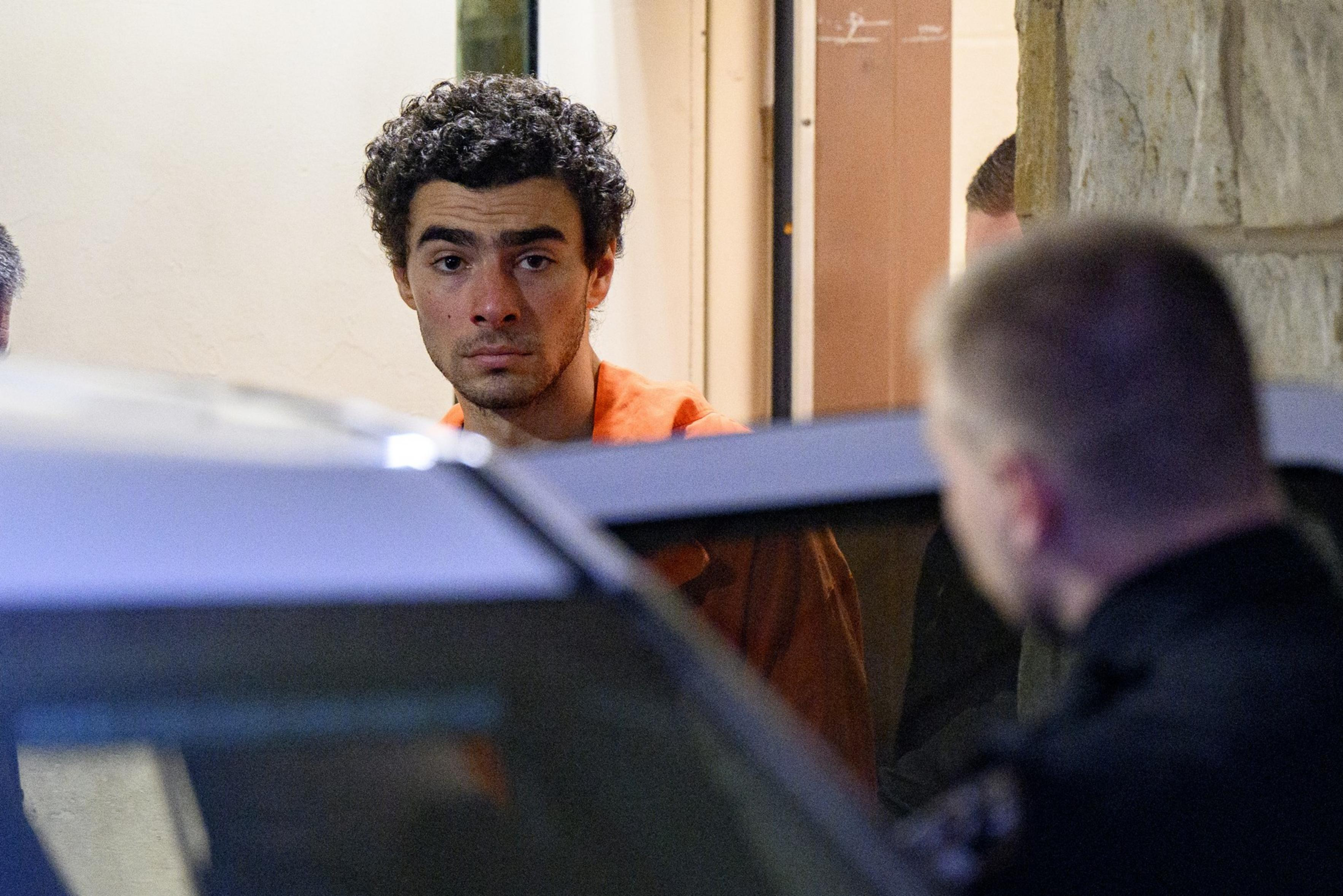 A man in an orange jumpsuit stares intently beside a vehicle, with an officer nearby, in a dimly lit setting.