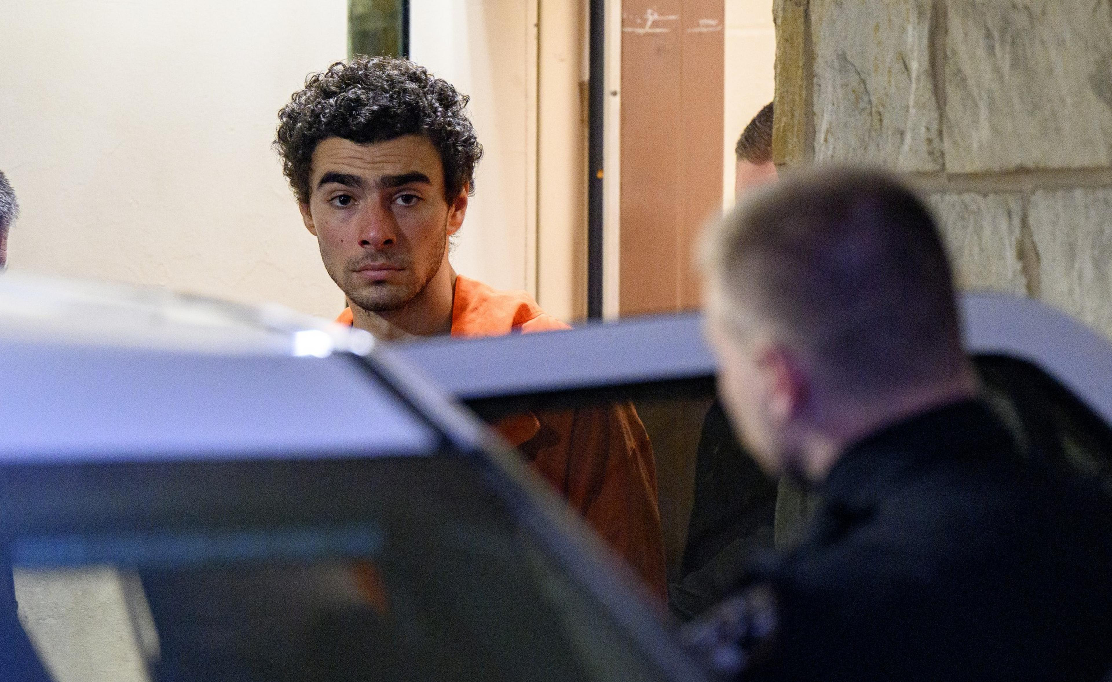 A man in an orange jumpsuit stares intently beside a vehicle, with an officer nearby, in a dimly lit setting.
