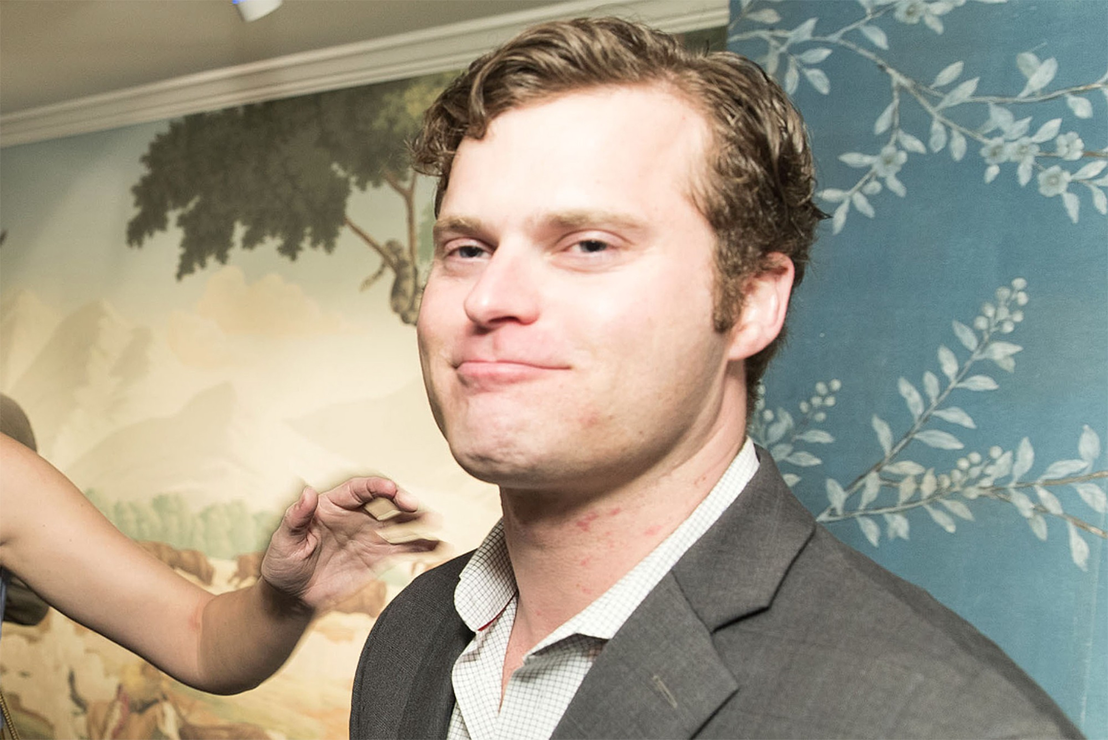 A man in a suit smirks while someone's hand makes a playful gesture near his face.