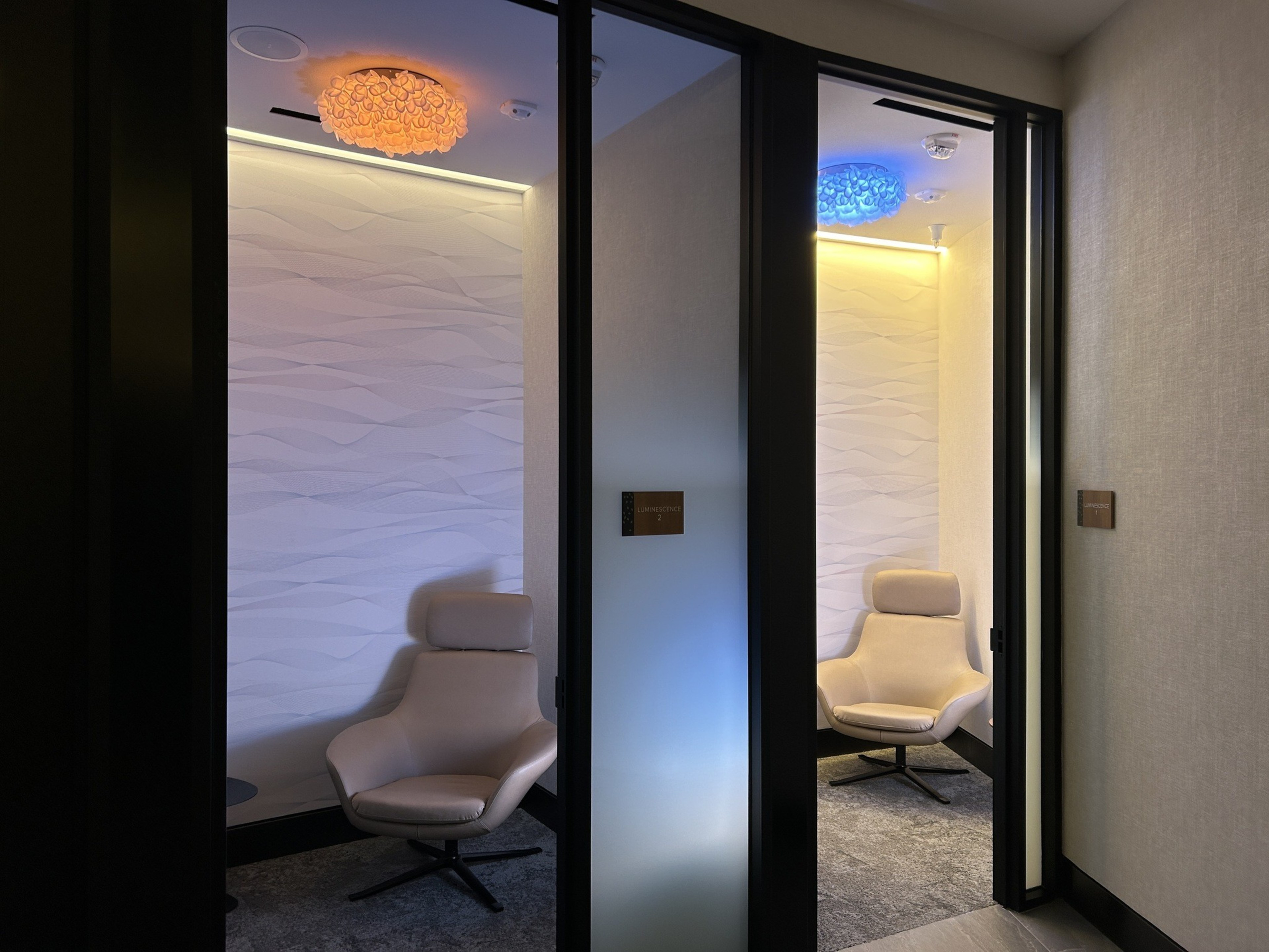 The image shows two small, modern rooms with armchairs, muted lighting, textured white walls, and colorful ceiling lights, one orange and one blue. Signs read &quot;LUMINESCENCE.&quot;