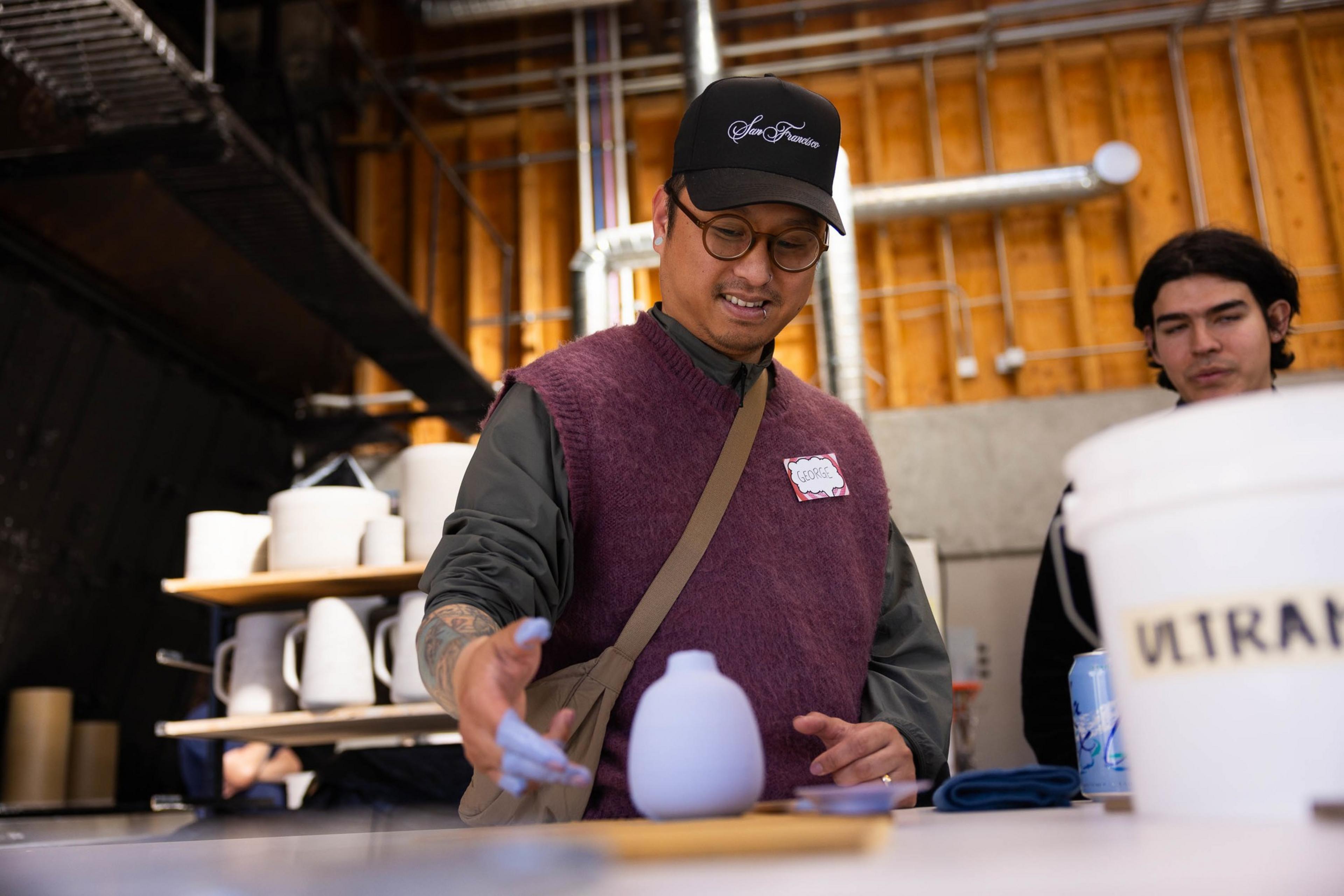 A man in a cap and vest attentively examines a small vase on a table, with an instructor and pottery nearby.