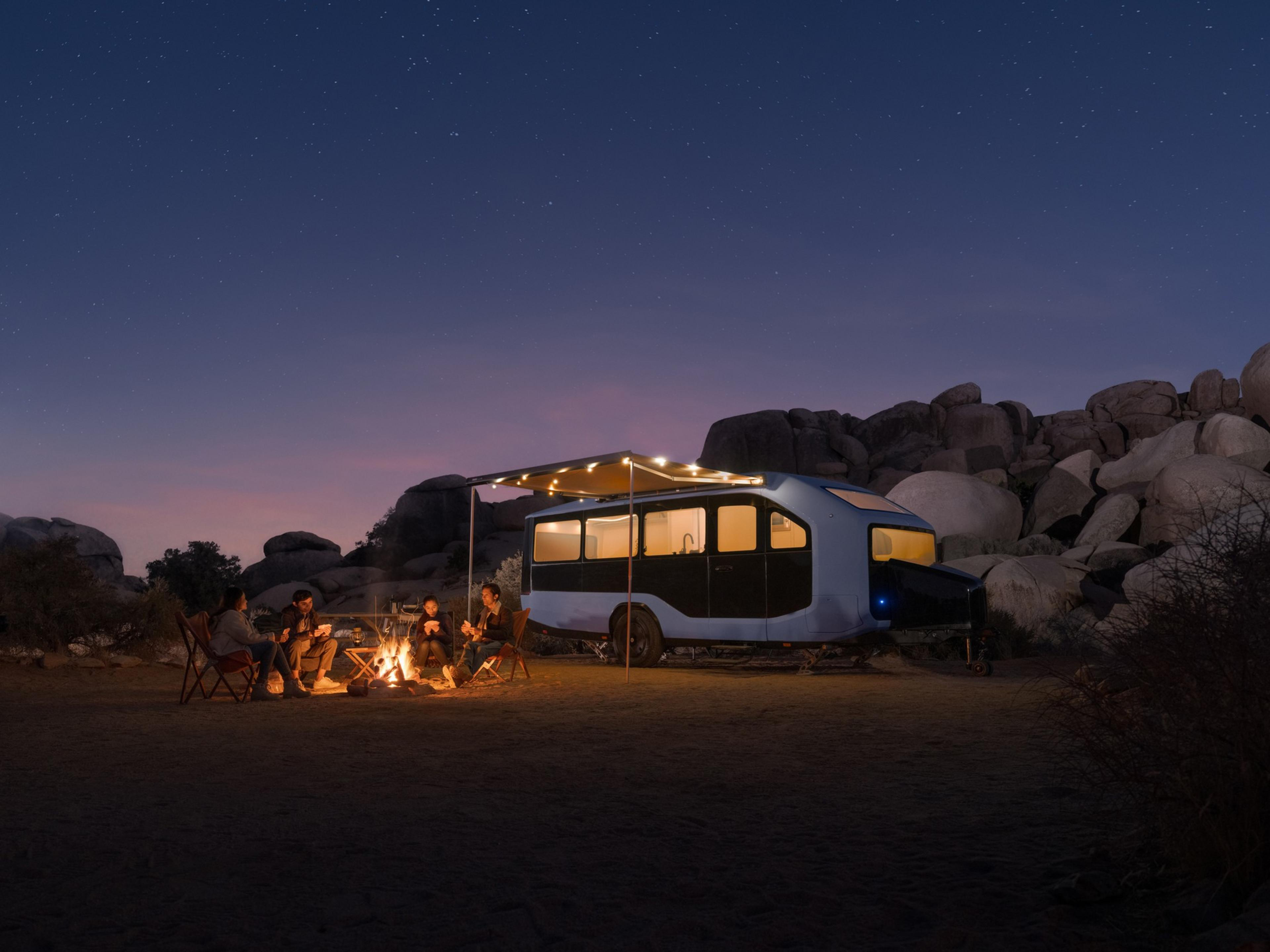 A group of people sit around a campfire at dusk, near a modern trailer with string lights, surrounded by boulders under a starry sky.