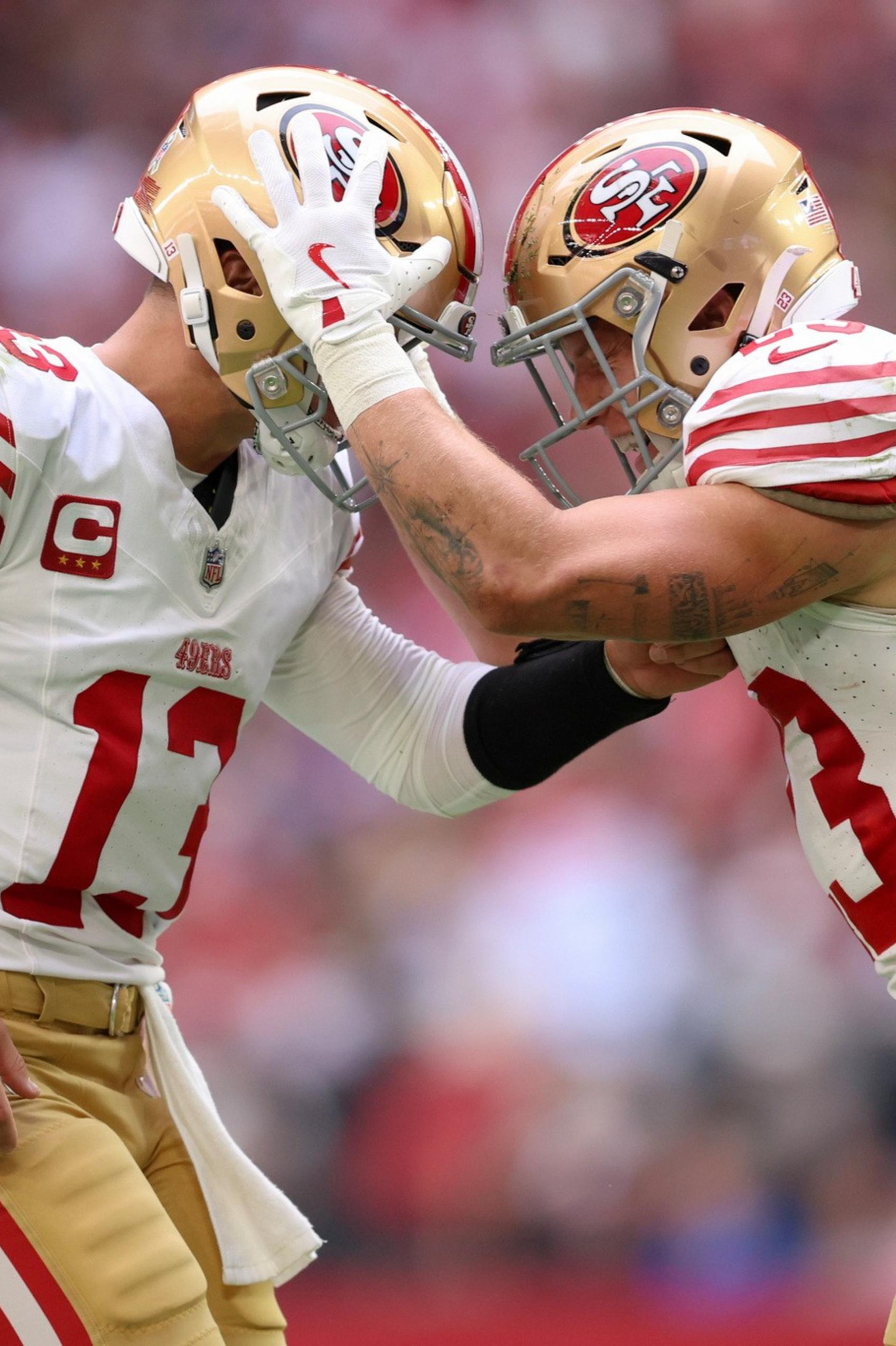 Two San Francisco 49ers football players in gold helmets and white jerseys touch helmets in a celebratory gesture during a game.