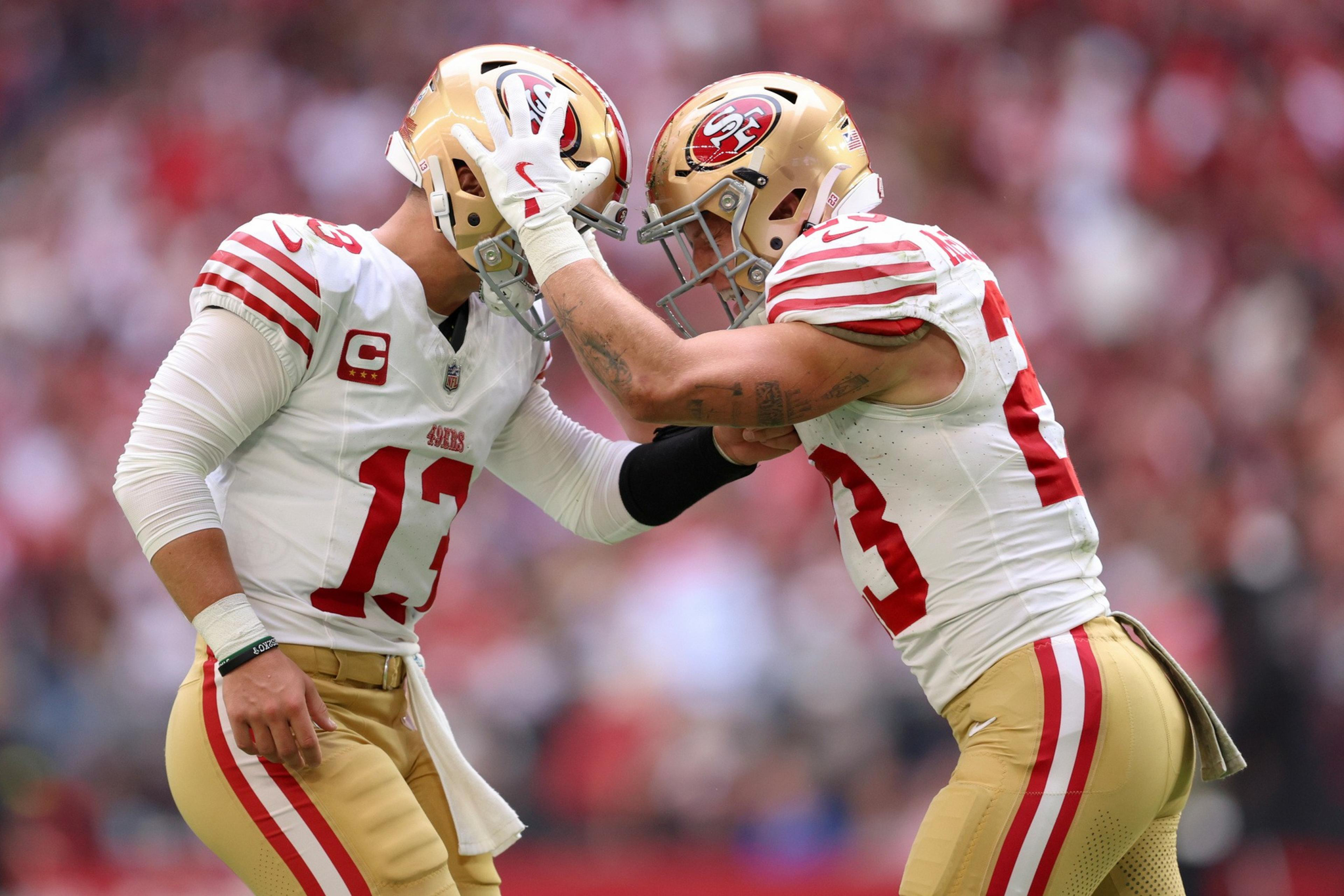Two San Francisco 49ers football players in gold helmets and white jerseys touch helmets in a celebratory gesture during a game.