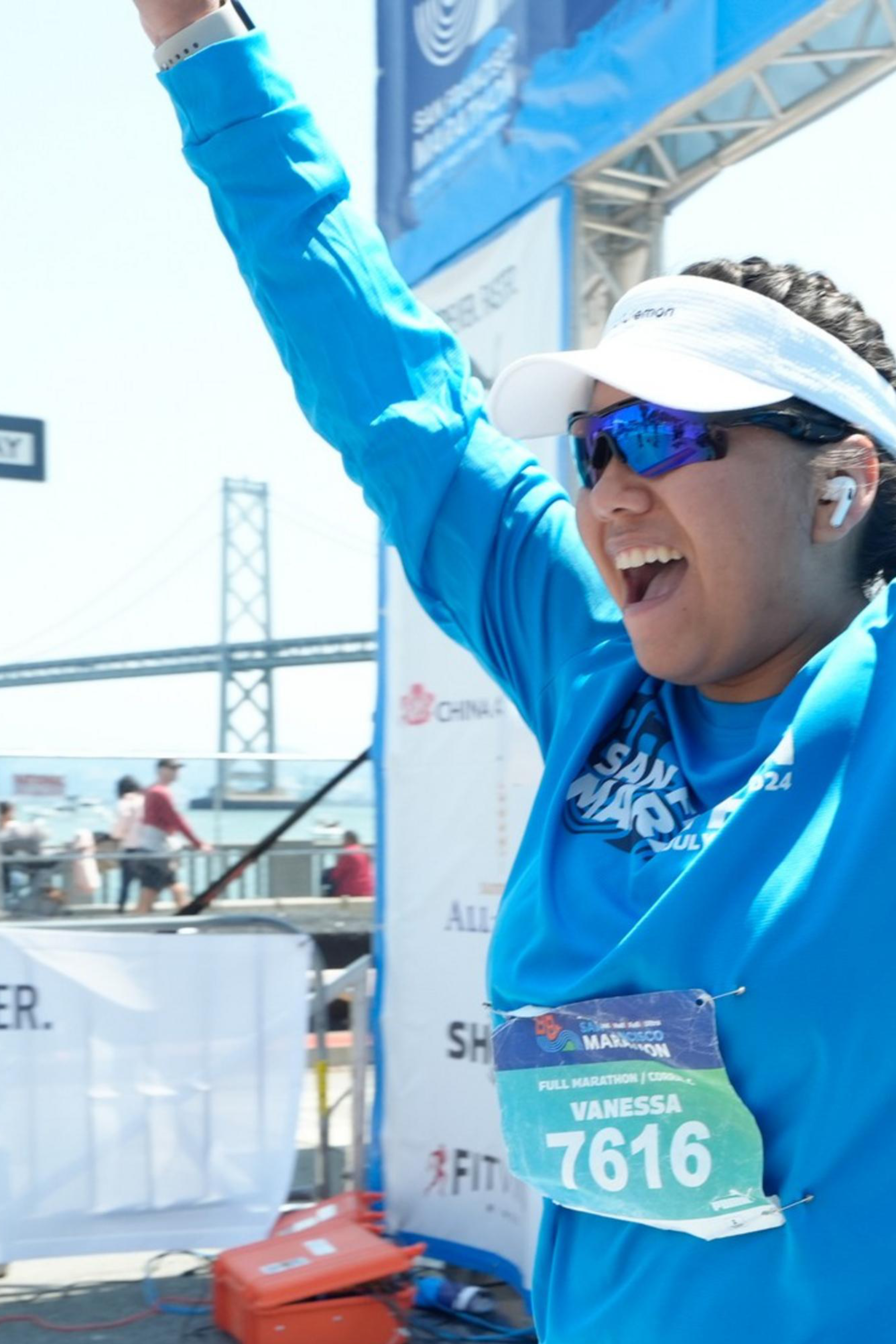 A joyful runner in a blue shirt, bib number 7616, and sunglasses, raises her arms in triumph at a marathon finish line, with a bridge and cheering spectators in the background.