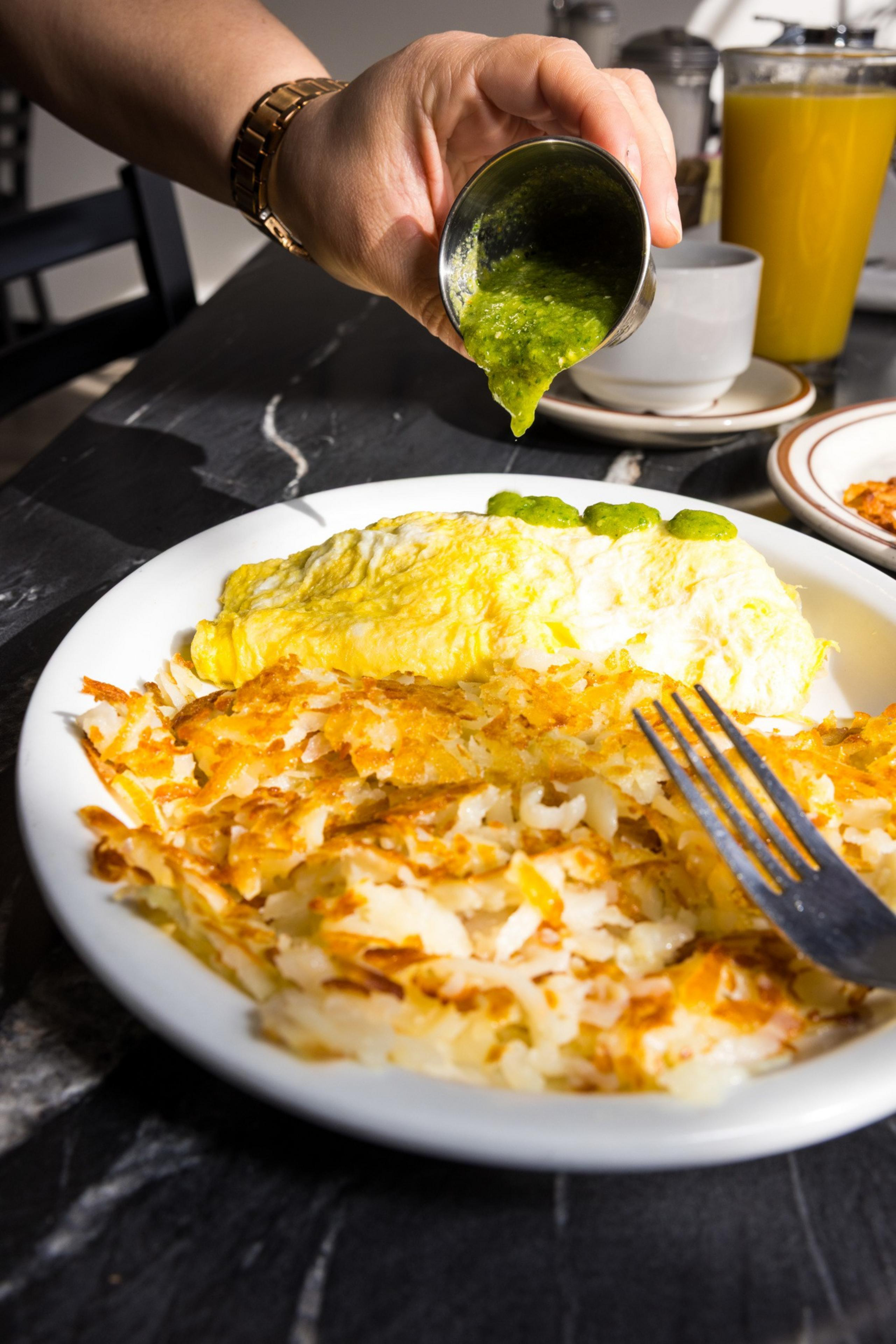 A hand pours green sauce over an omelet on a plate with crispy hash browns. A fork rests on the plate, and orange juice and a cup sit in the background.