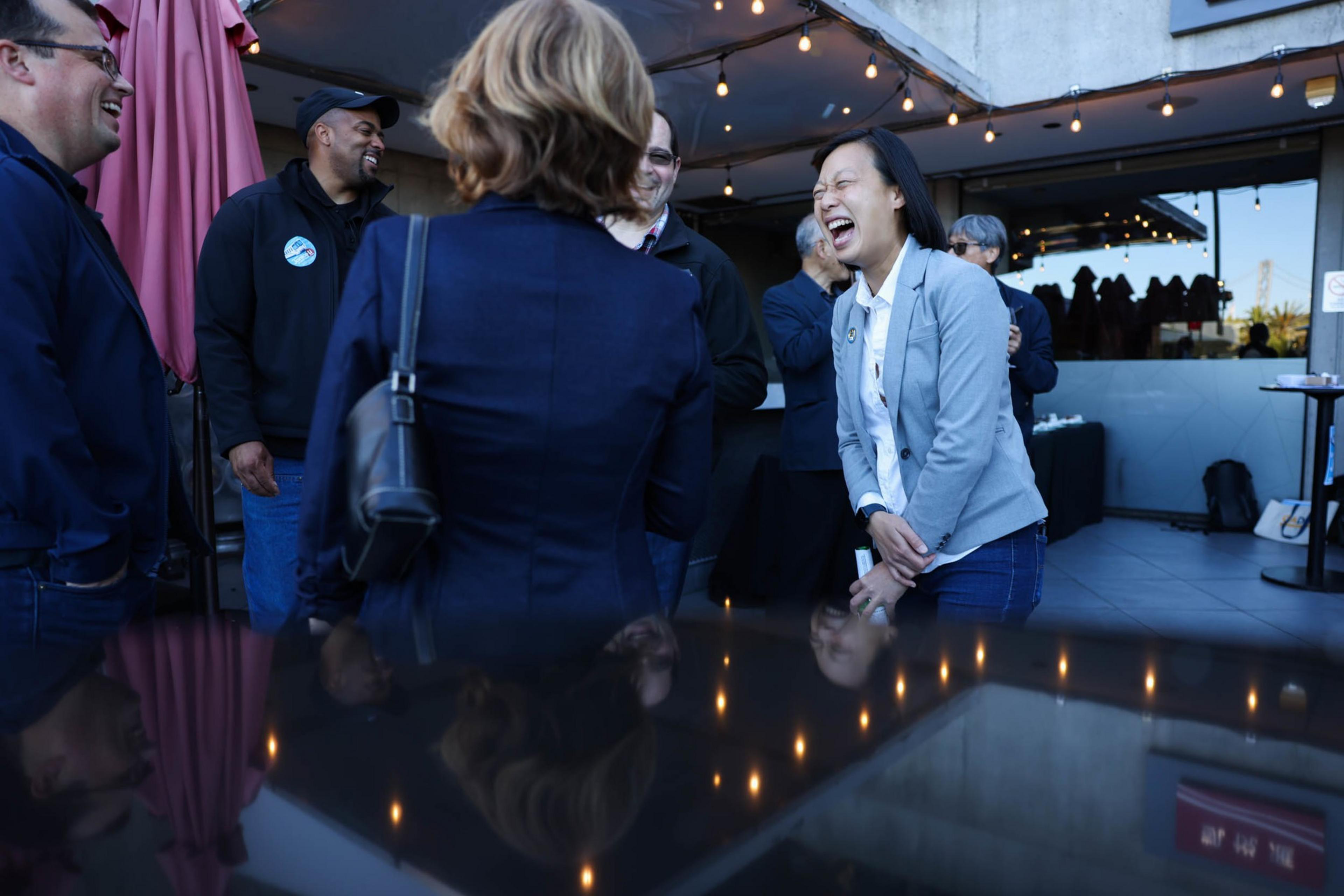 A group of people stand outdoors under string lights, laughing and talking, with one woman in a light jacket caught in a hearty laugh.