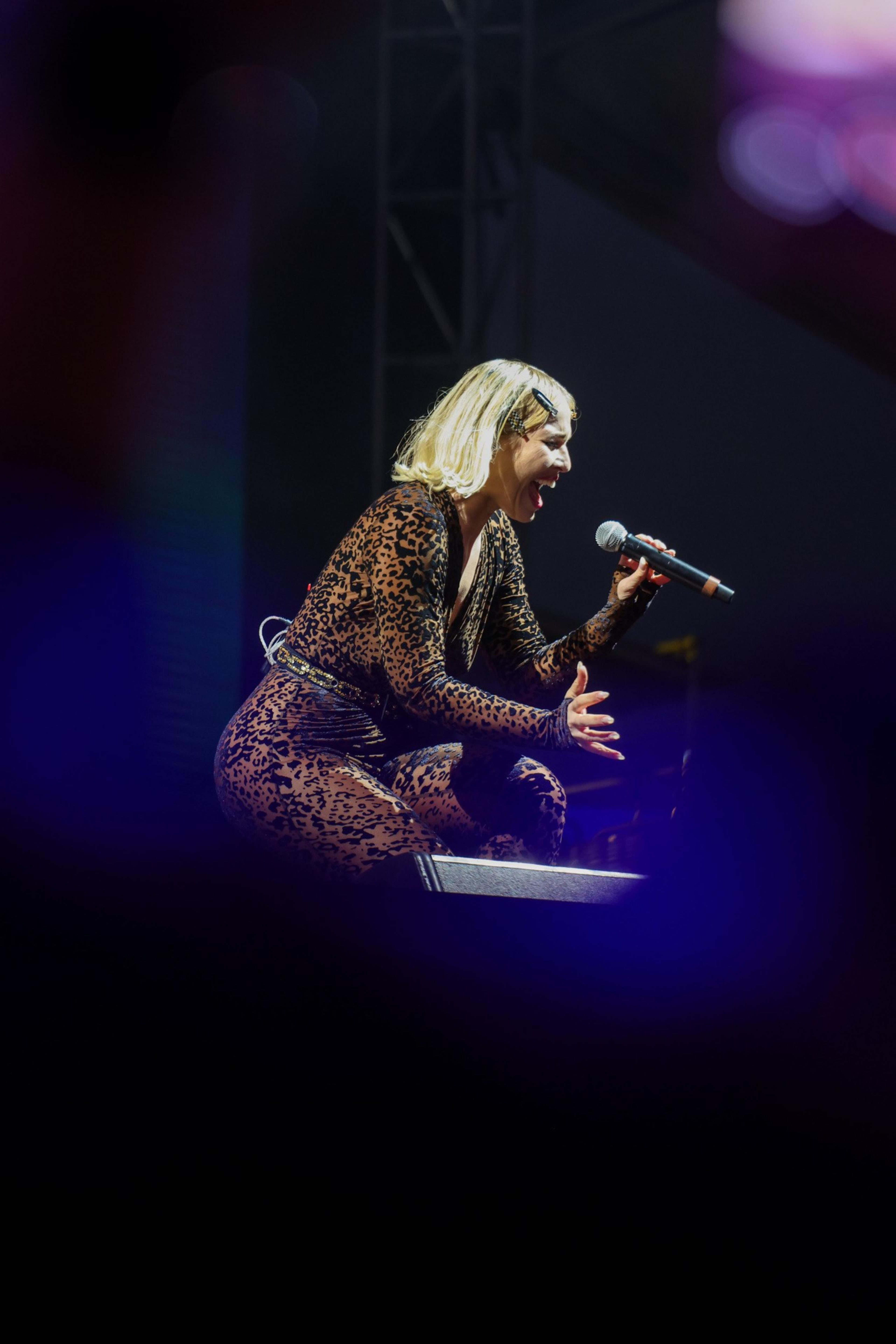 The image features a performer in a leopard print outfit, passionately singing into a microphone on stage with dramatic lighting and a dark background.