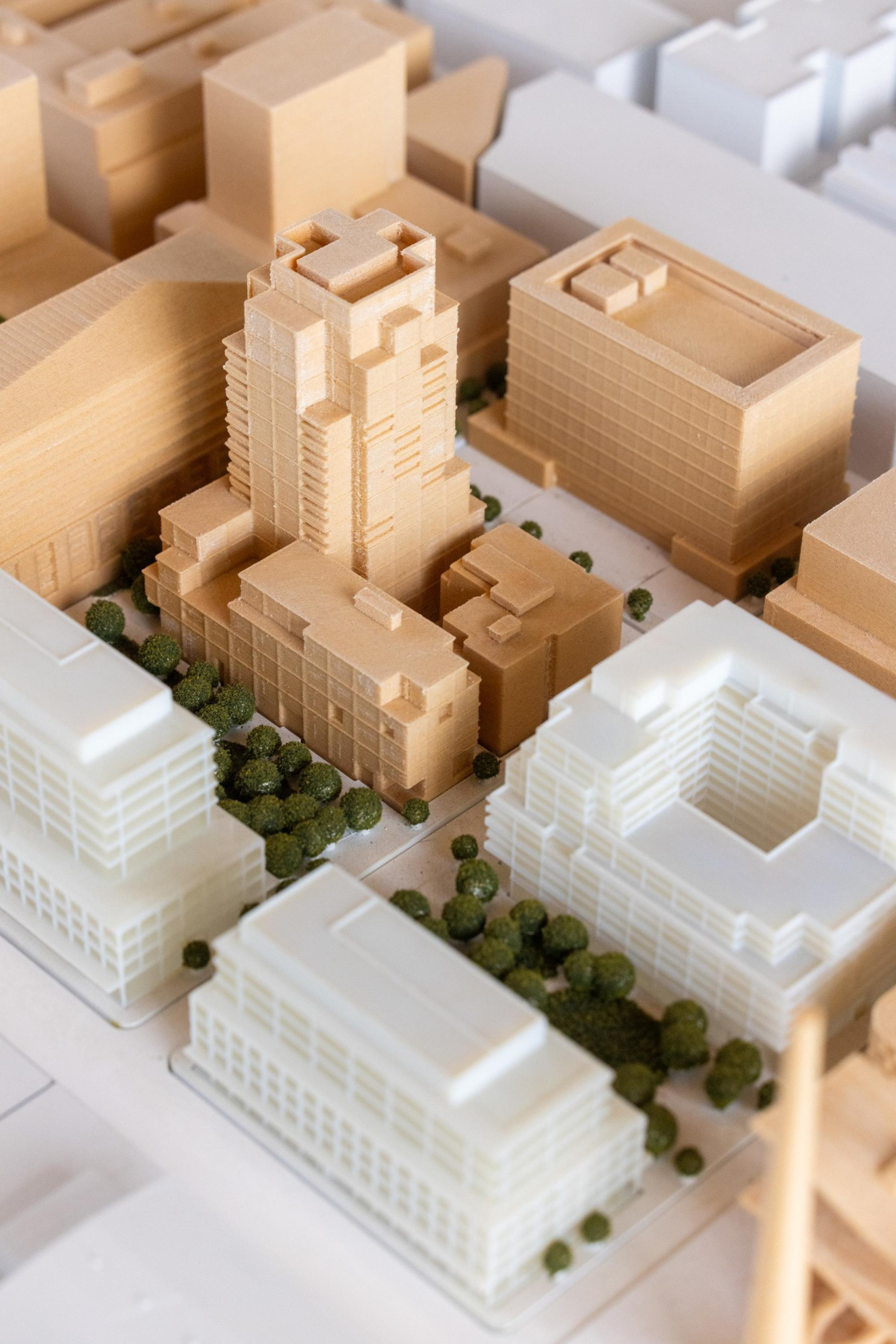 A detailed architectural model of an urban area features multiple buildings, with a tall, intricately designed central tower surrounded by smaller structures and green trees.