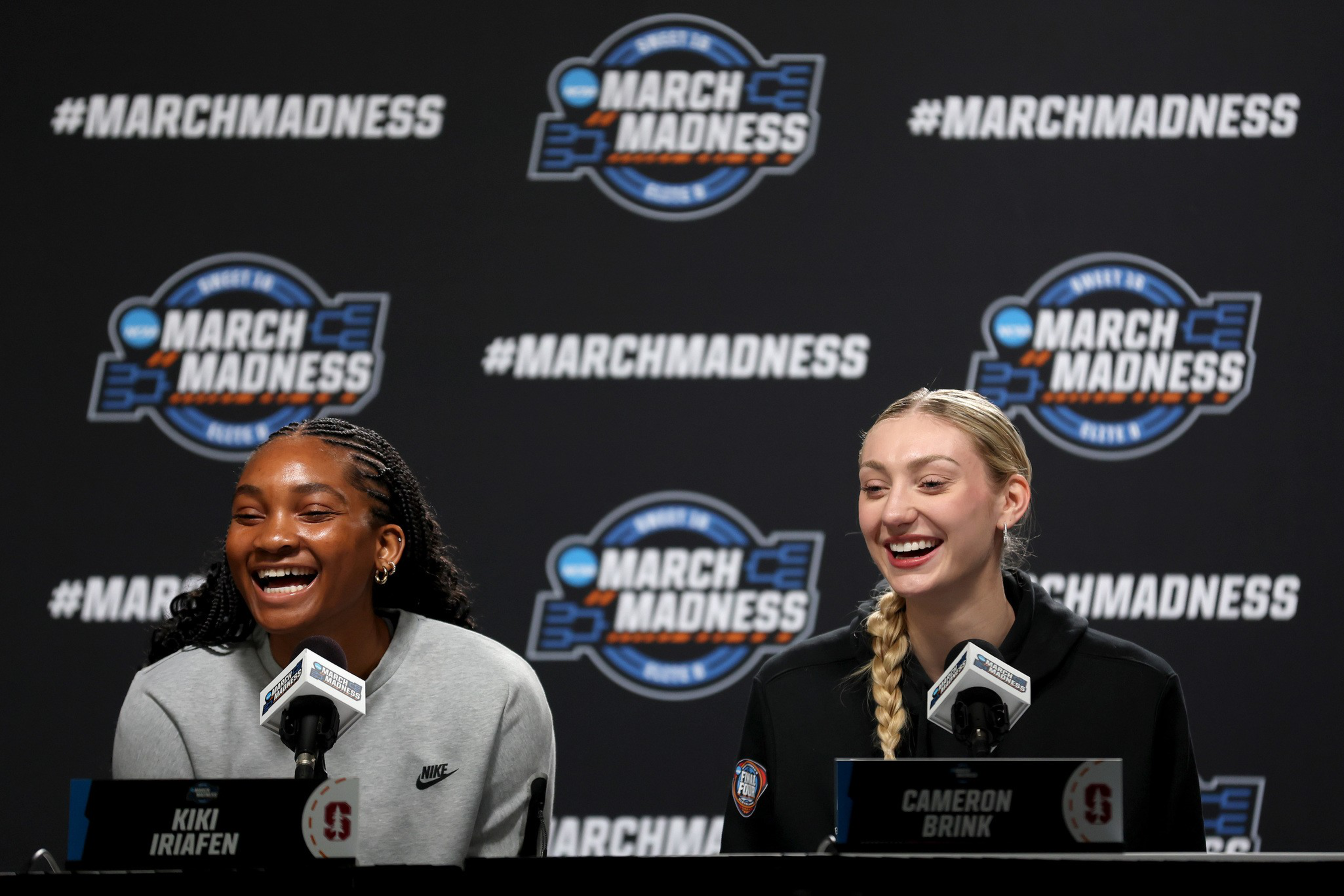 Two women, Kiki Iriafen and Cameron Brink, smile and speak into microphones at a March Madness press conference with a branded backdrop.