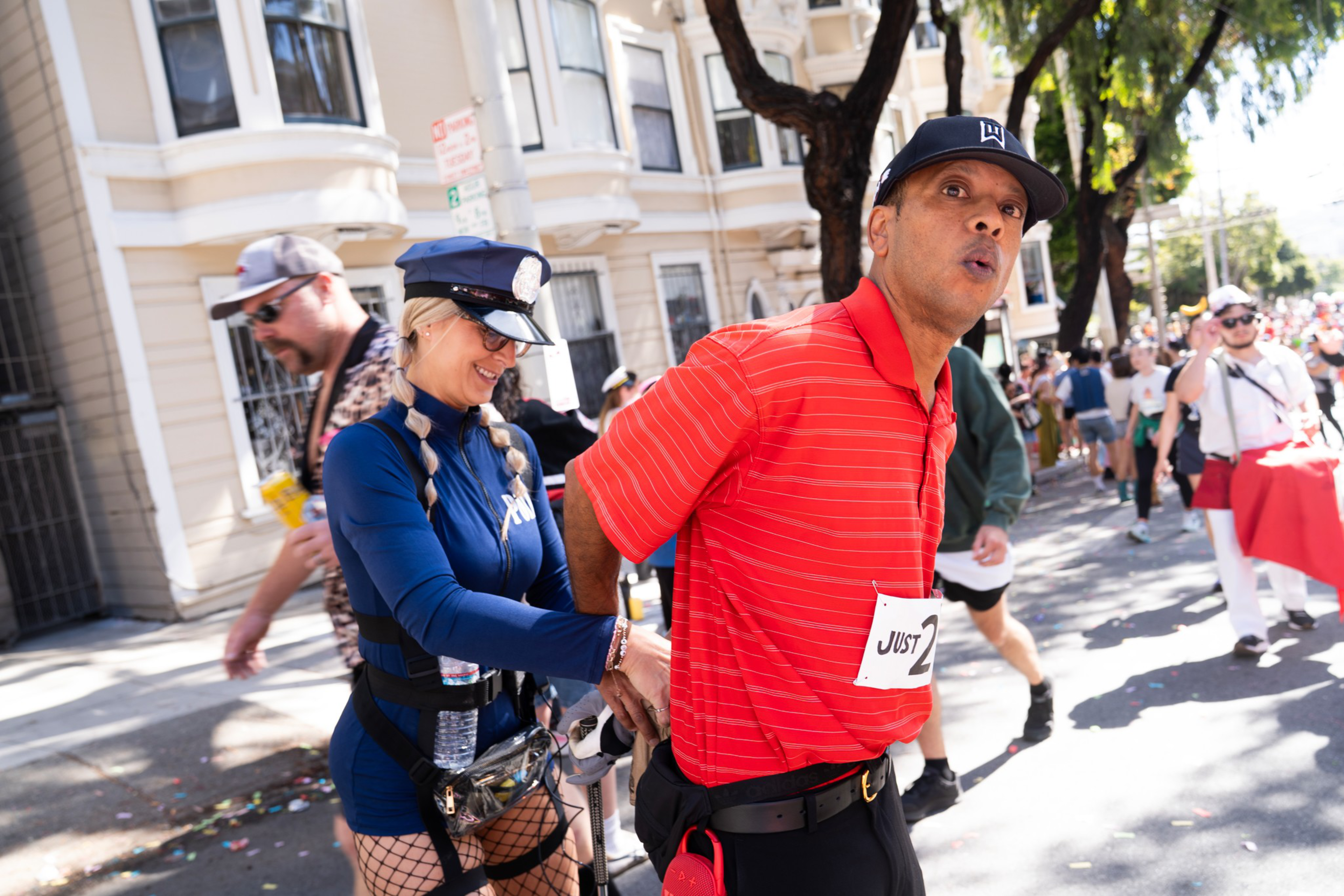 A person dressed as a police officer playfully handcuffs a man in a red shirt and black cap during an outdoor event with people and buildings in the background.