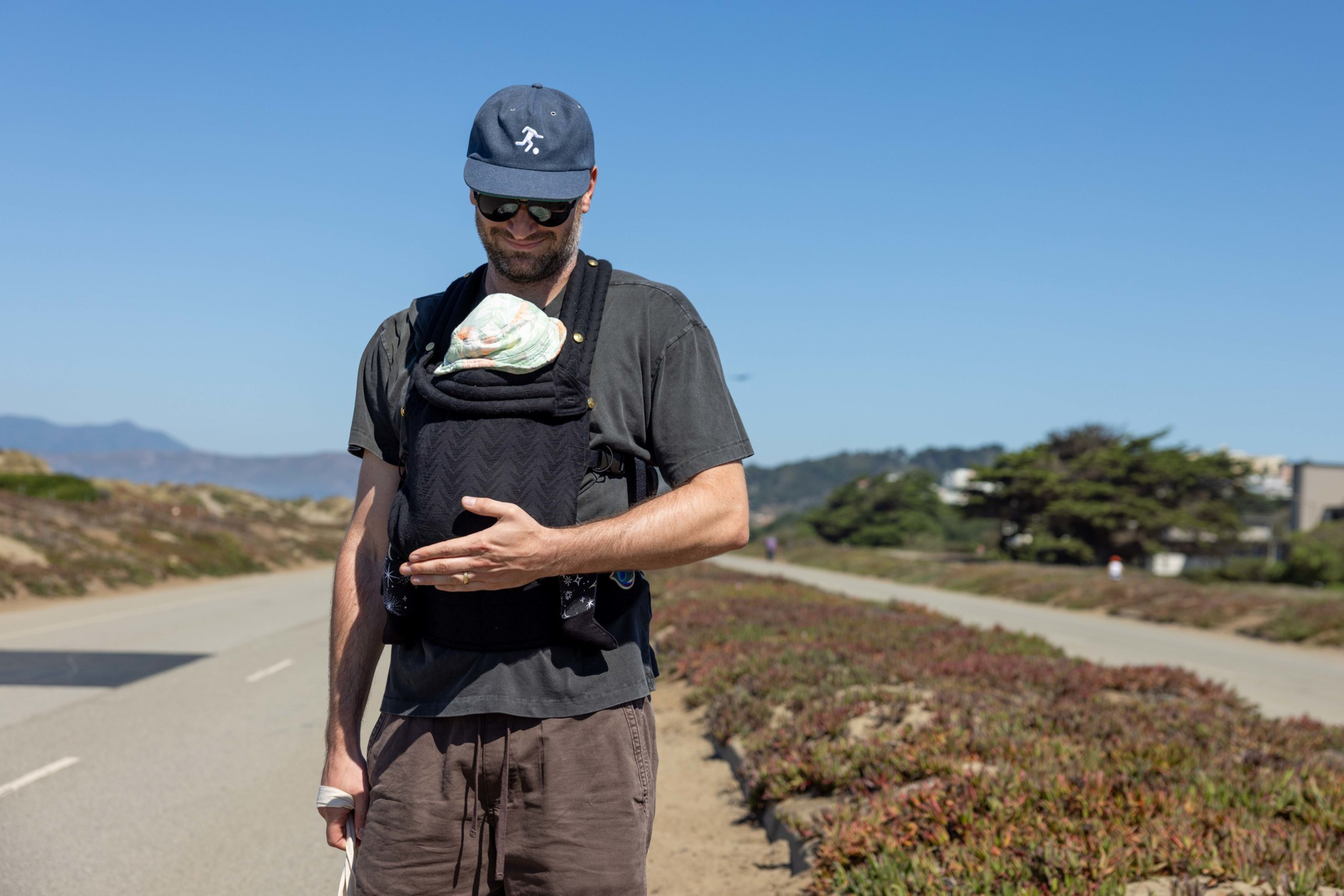 A man wearing sunglasses and a baseball cap walks outside on a path carrying a baby in a black baby carrier on his chest.