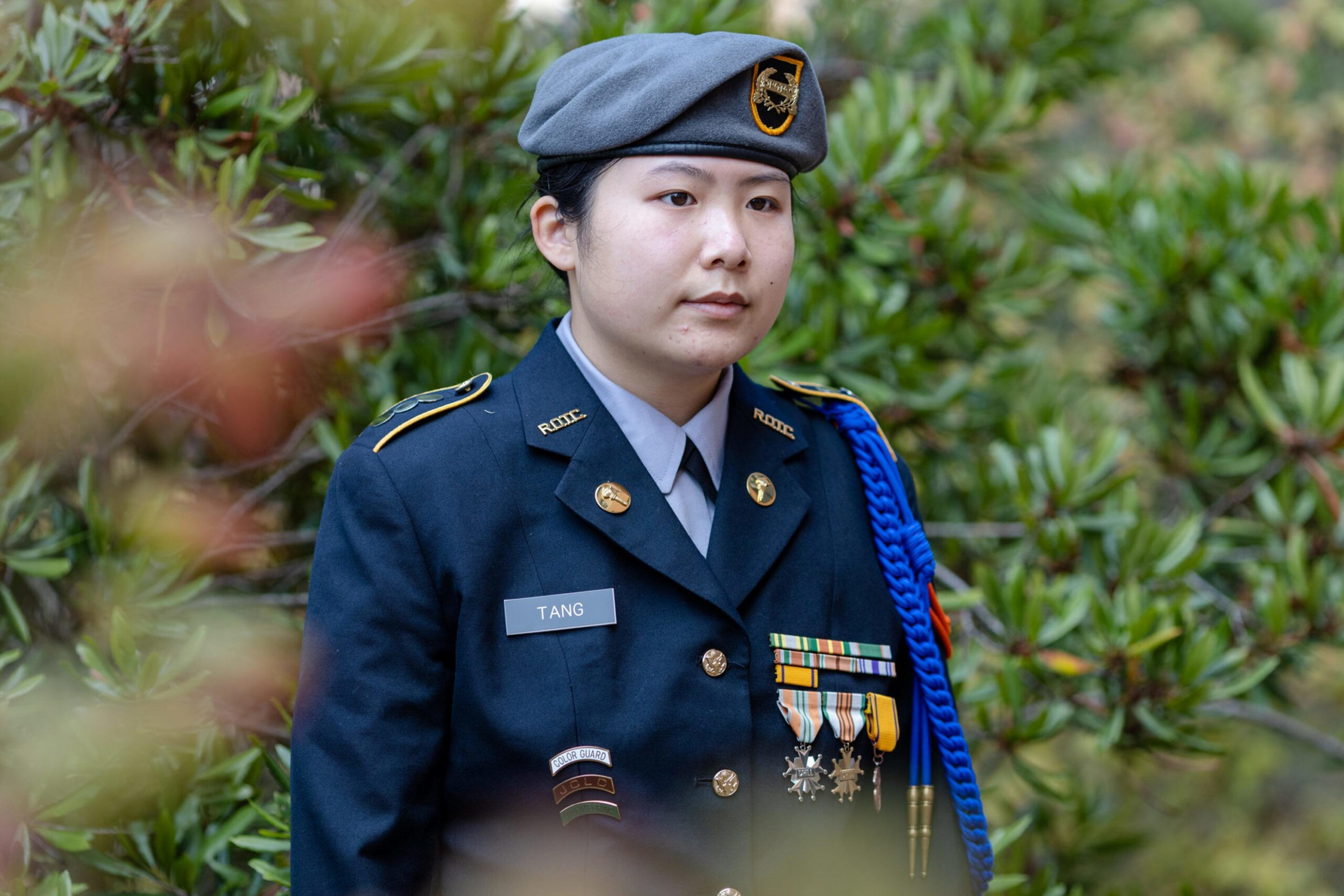 A person in a formal military uniform with the name tag "Tang," wearing a gray beret and decorated with medals and ribbons, stands outdoors near greenery.