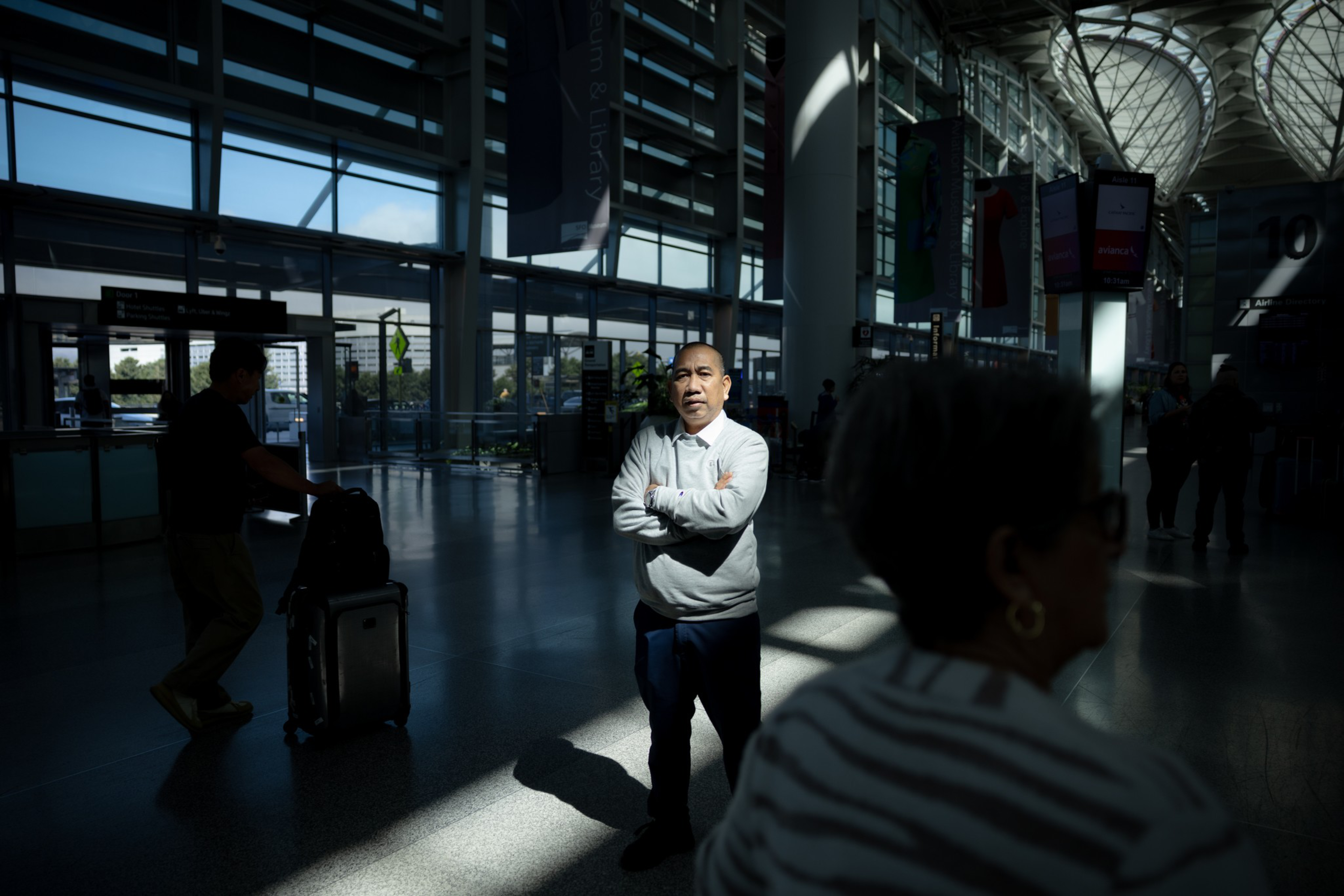 A man in a light sweater stands with crossed arms in a sunlit spot inside a large, modern airport terminal with glass walls and people around.