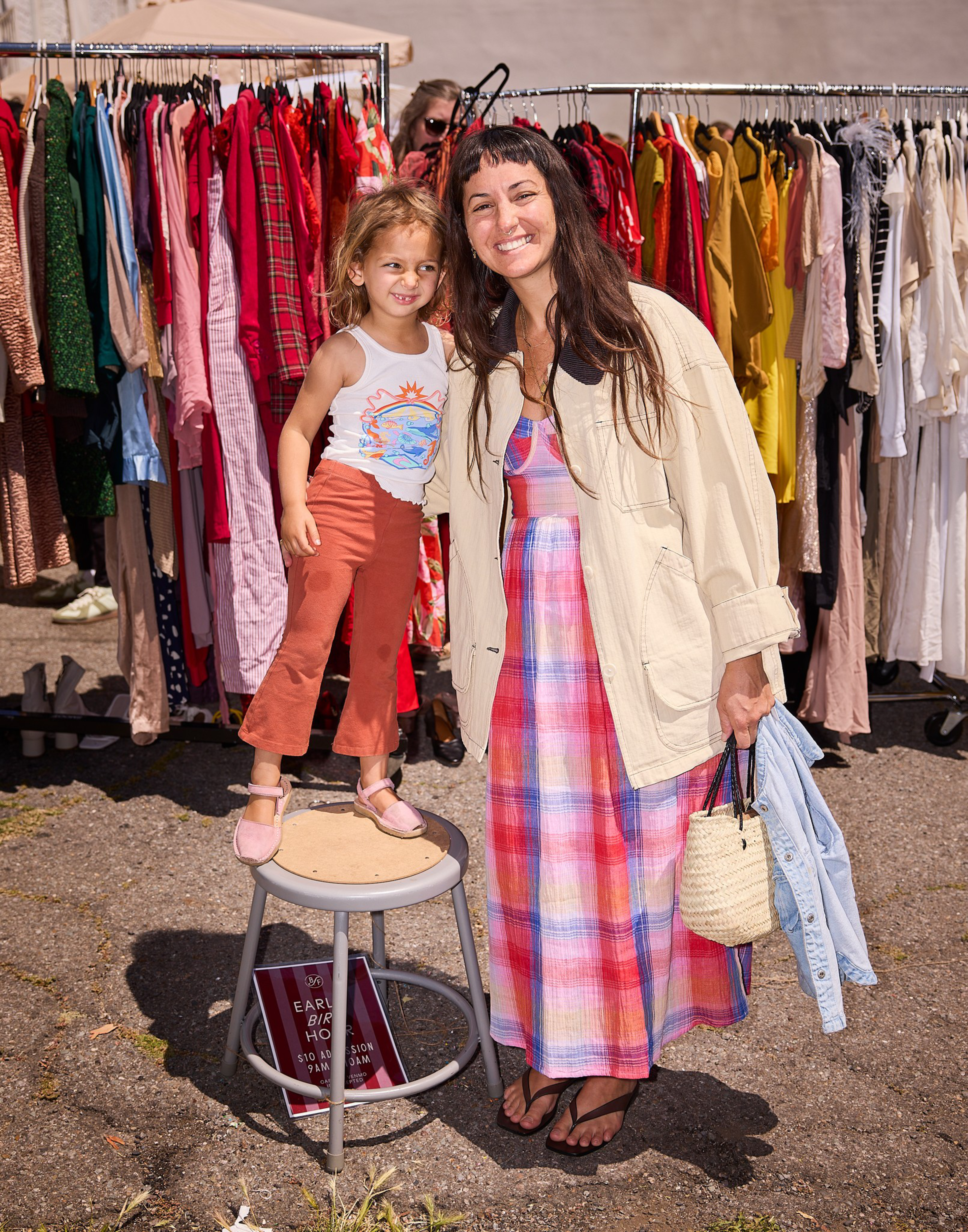 A smiling woman in a colorful plaid dress and cream jacket stands next to a young girl in a white tank top and rust pants, both in front of a clothing rack.
