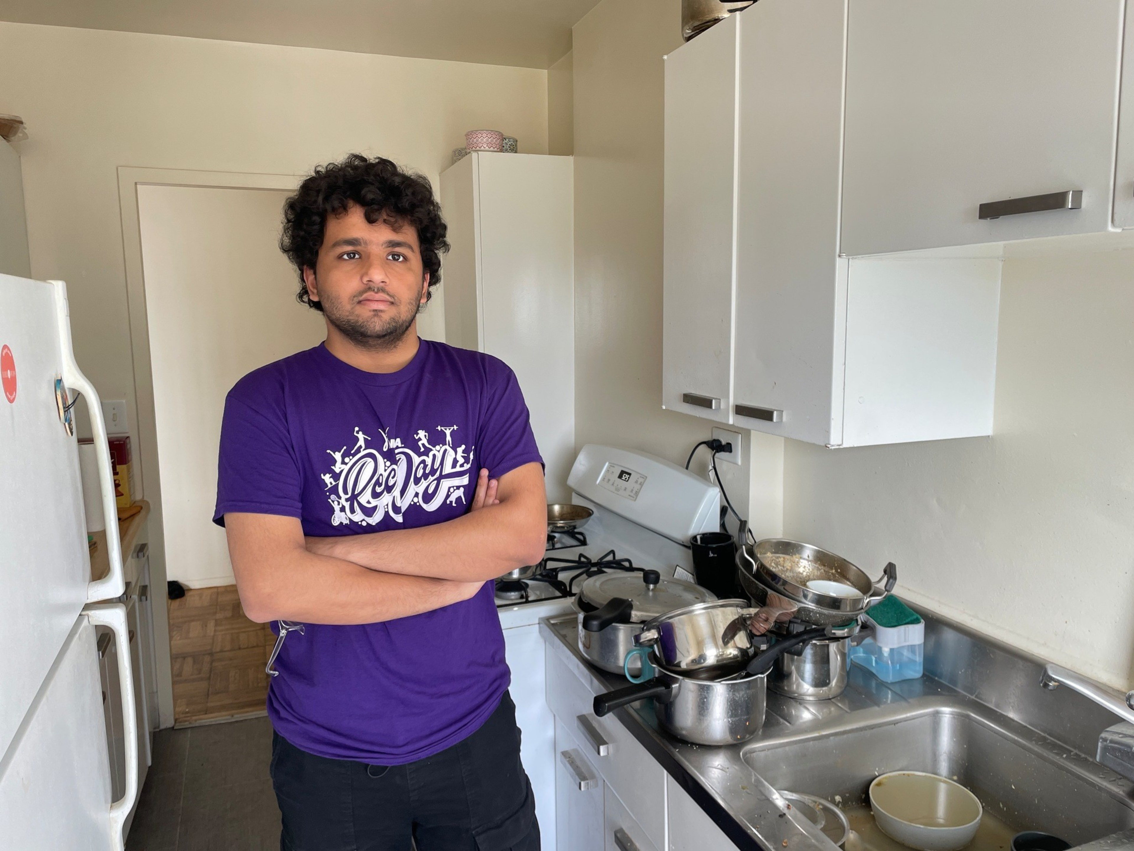 A man in a purple shirt stands with arms crossed in a kitchen with white cabinets, a stove, and a cluttered countertop filled with pots and a sink with dishes.