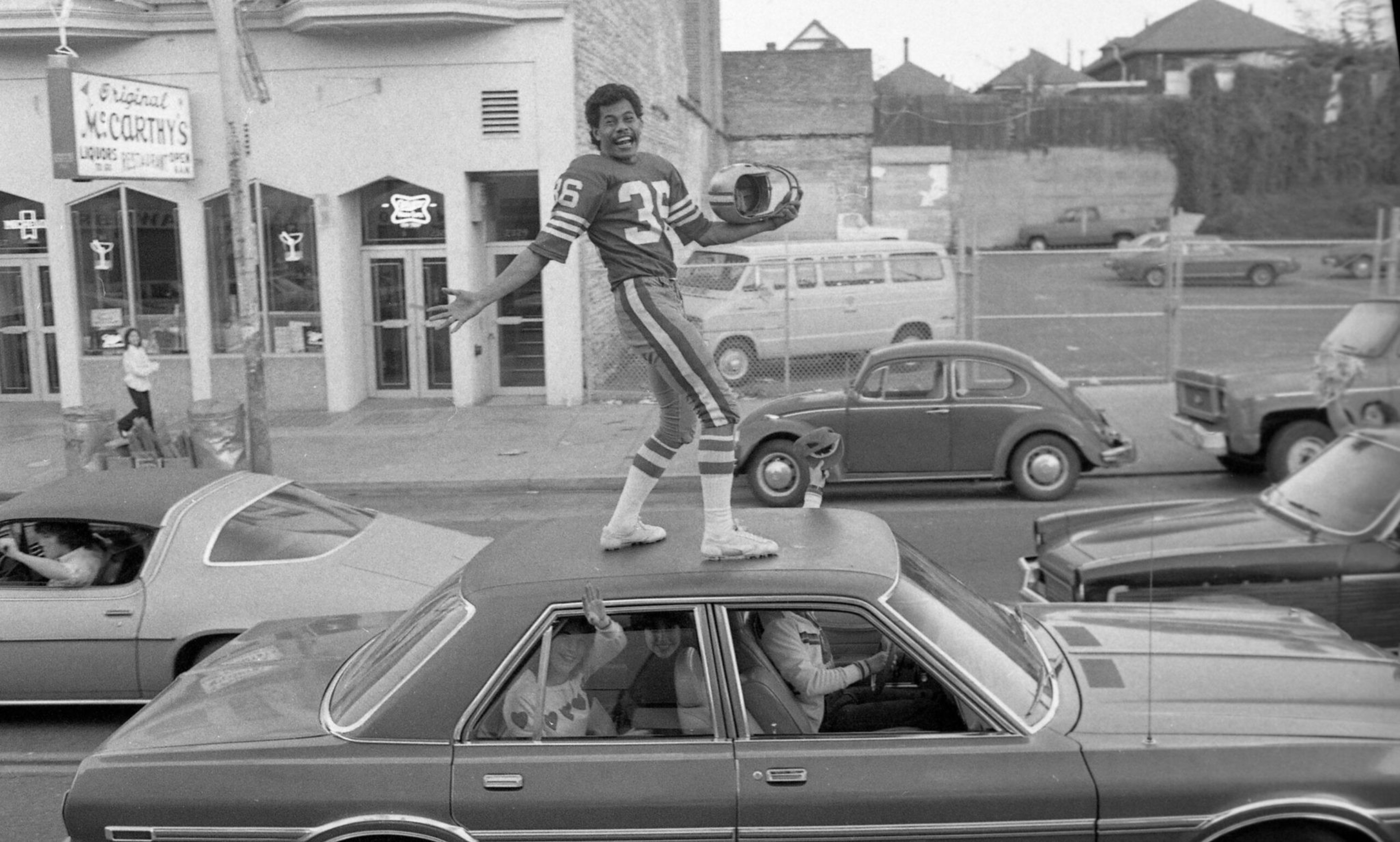 A man in a football uniform stands on the roof of a car, holding a helmet and smiling, while people inside the car look out the windows.