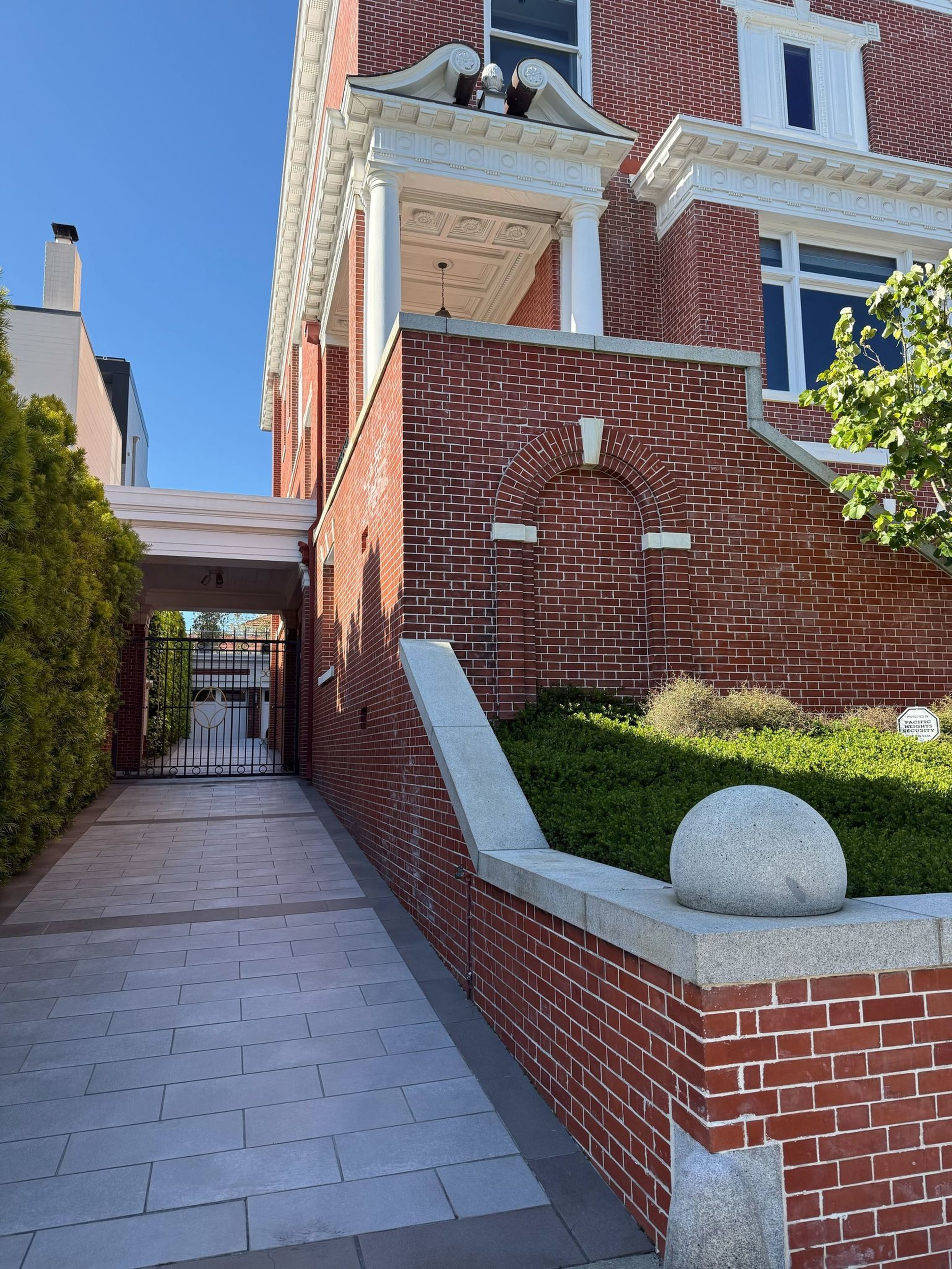 A red brick building with white columns and detailed cornices, a tiled walkway alongside it, green bushes, and a gated entrance at the end of the path.