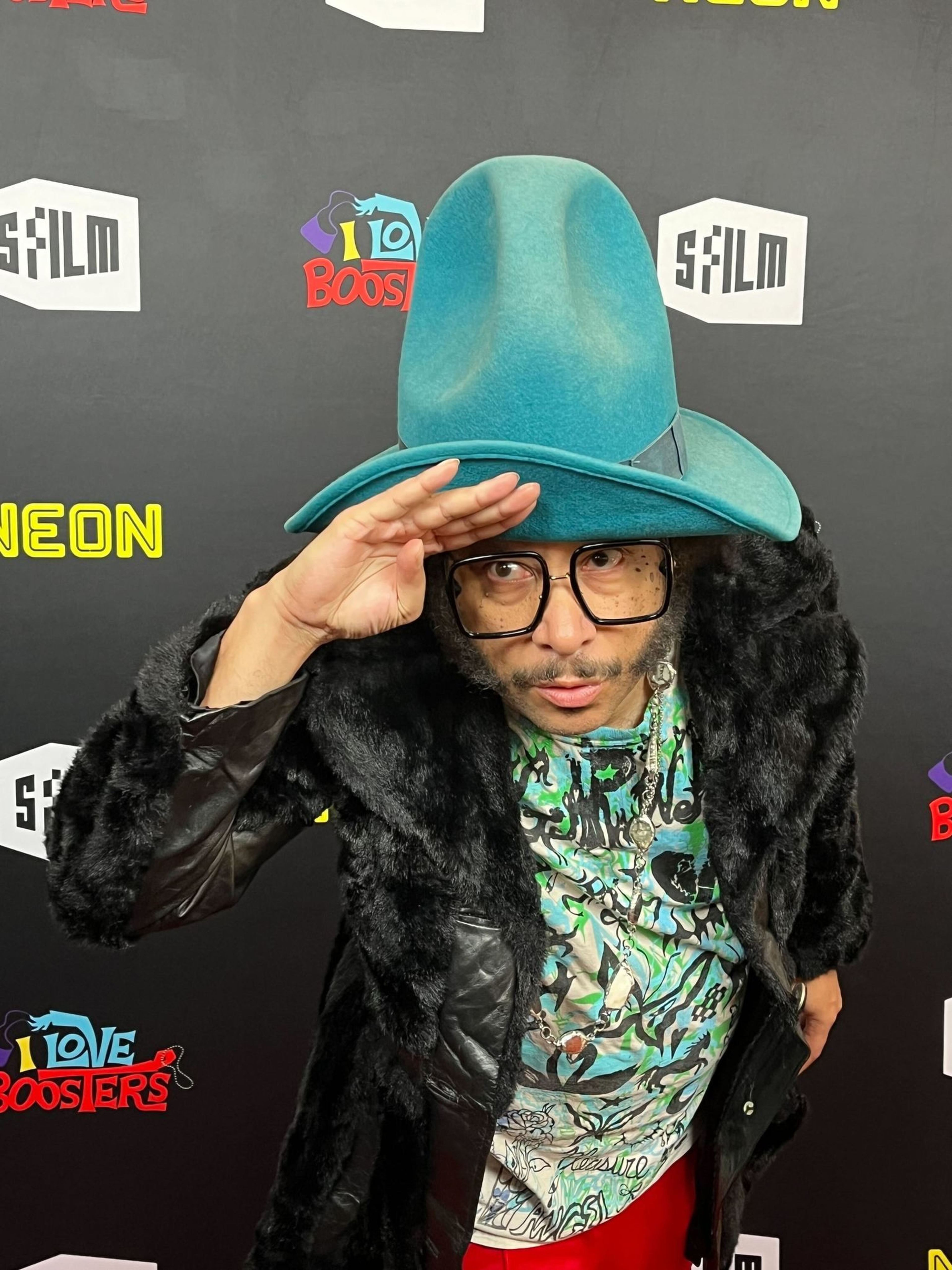 A person wearing a large teal hat, oversized black glasses, a patterned shirt, and a black fur coat poses with a hand salute against a branded backdrop.