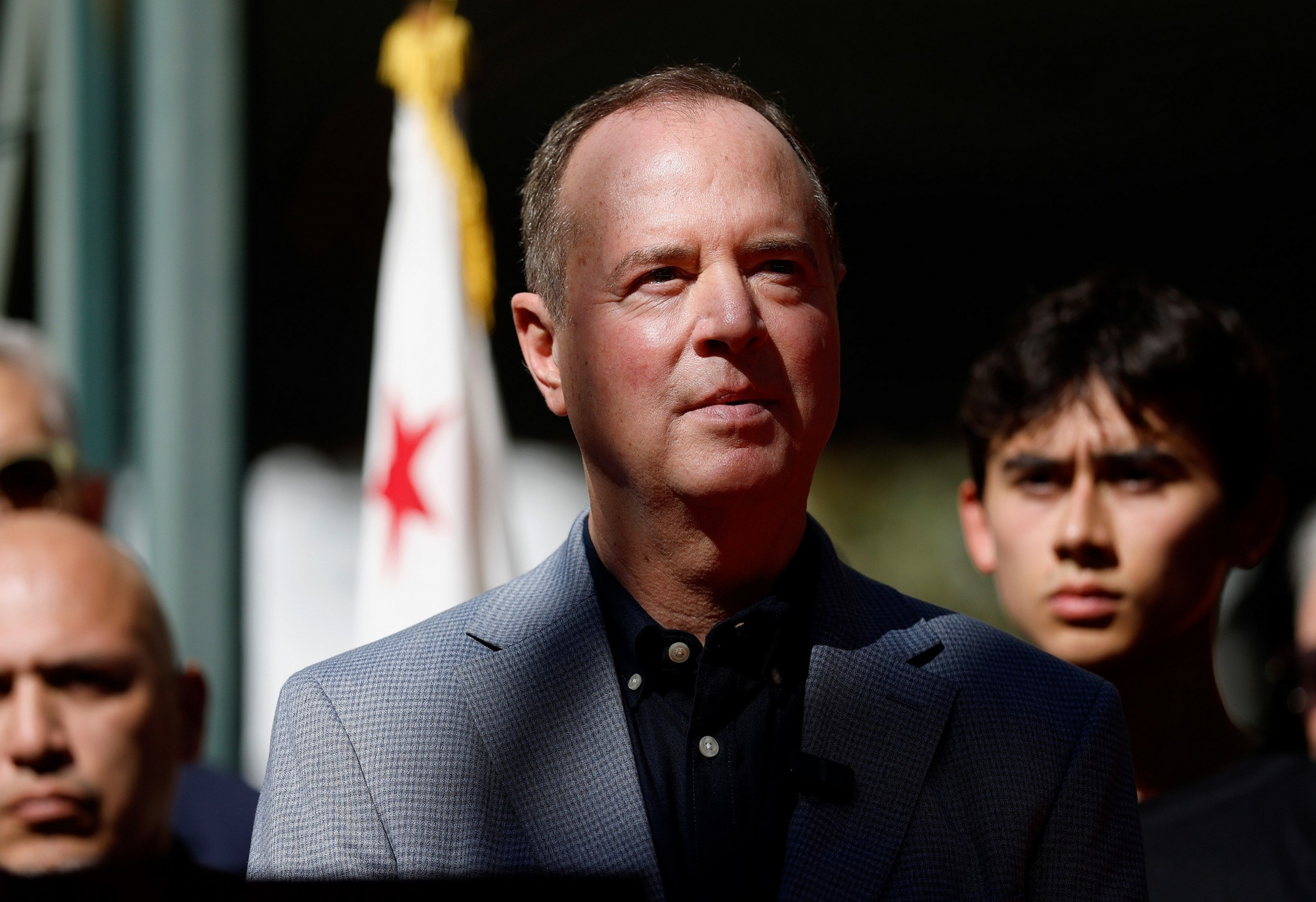 A man in a gray blazer and black shirt stands in sunlight with a blurred California flag behind him and two serious-faced men in shadow.
