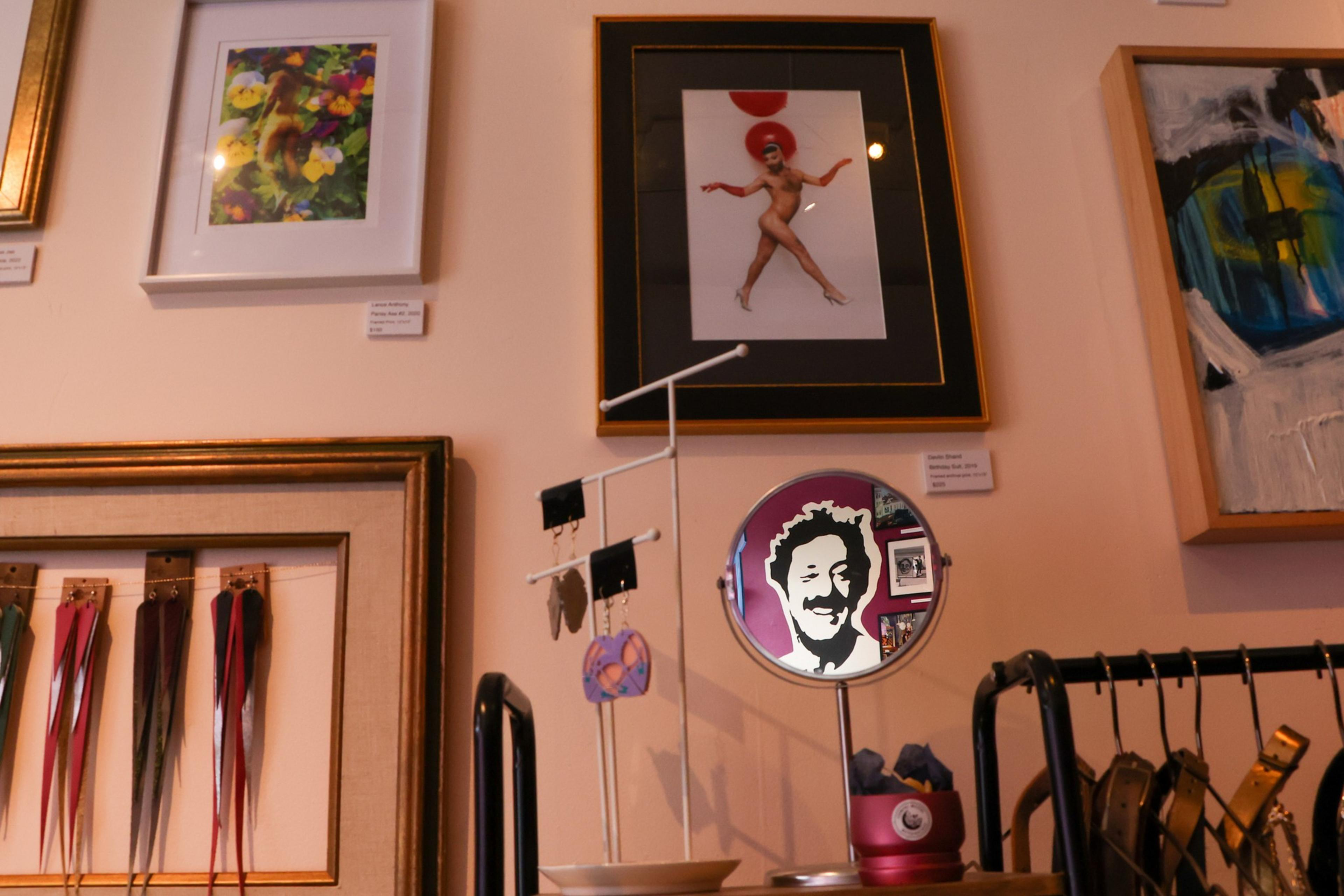 A gallery wall with framed colorful artworks, a circular mirror reflecting a smiling face, and a display table holding earrings and leather belts.